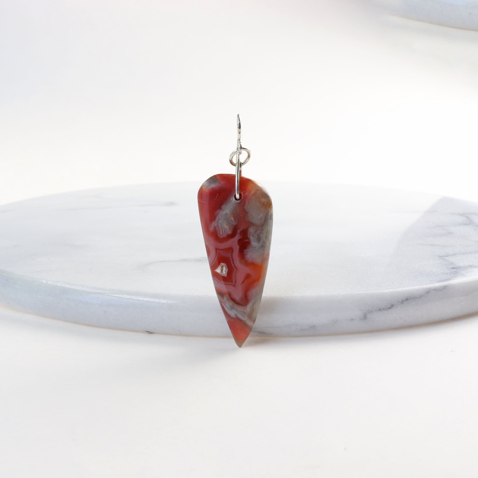 A Crazy Lace Agate arrow shaped gemstone pendant. Shown against a background of white marble.  The pendant has been handcut and freeform polished on both sides to reveal the unique swirling patterns and vibrant mauve deep reds and gray hues. Wear with complementary chain included or with you own favorite necklace.