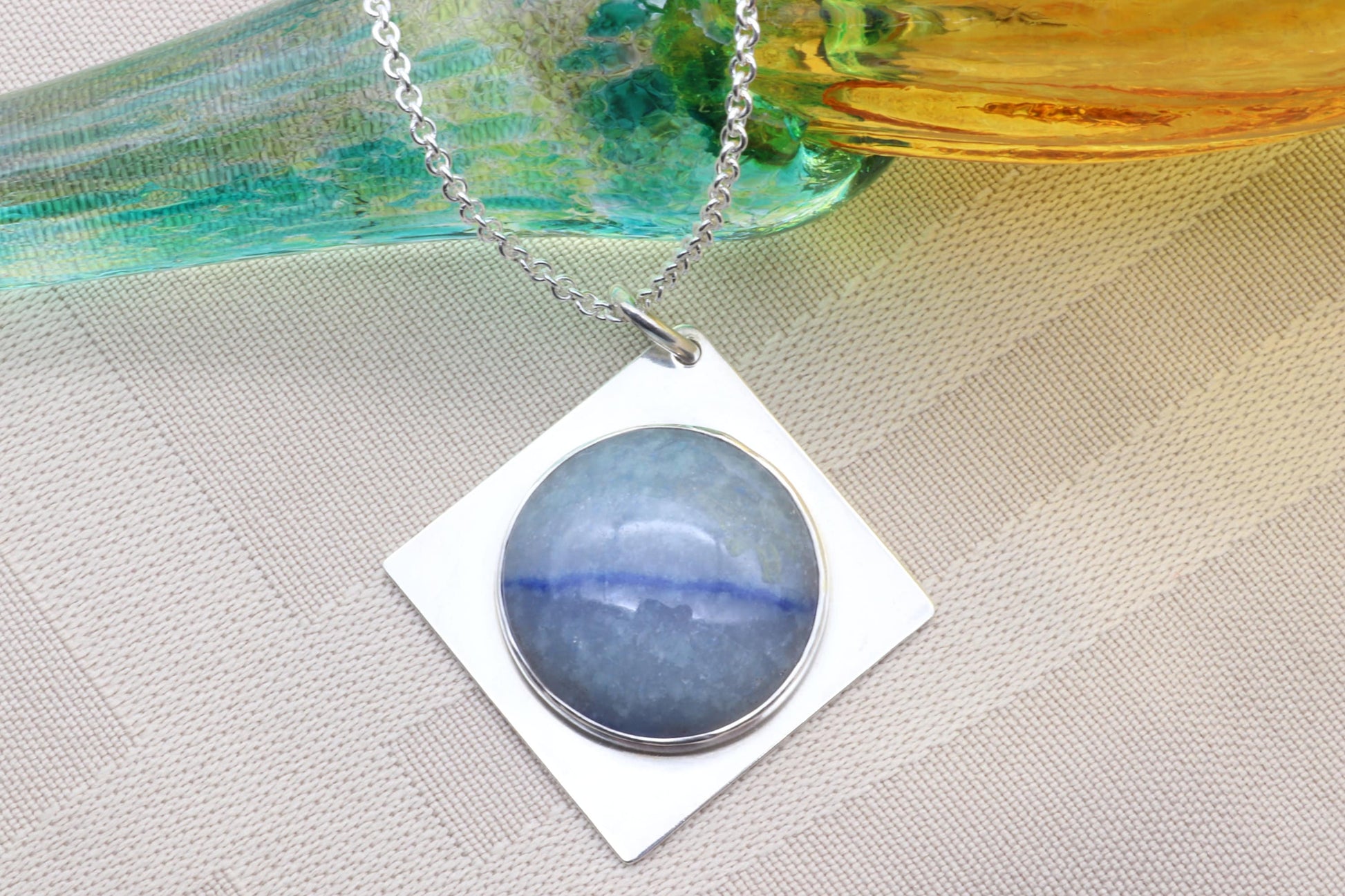 A close-up of a Celestial Blue Aventurine pendant with Saturn-like inclusions on a beige linen background leaning on a glass cylinder.