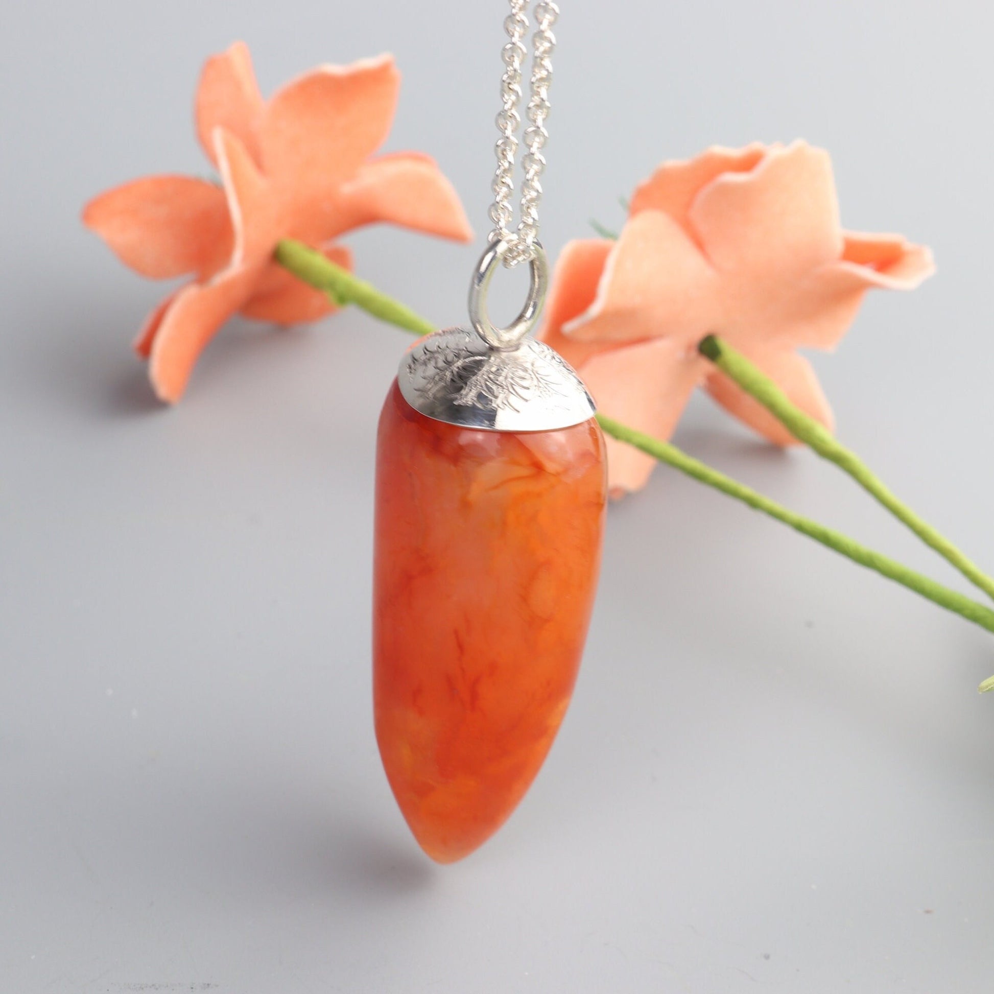 one-of-a-kind  2.15 inch Carnelian Crystal Agate pendant, hand-cut and freeform polished into a unique pepper-shaped form, reminiscent of a small orange sweet pepper, to highlight its vibrant natural colors and patterns.  Suspended from a silver chain against a white background.
