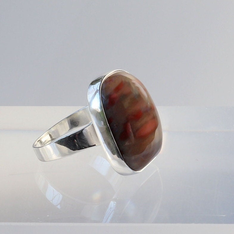Closeup of a handmade Petrified Rainbow Wood men's gemstone ring.  This gemstone has been handcut and polished in our Missouri studio to showcase the natural red and brown hues and captivating patterns.  Encased in sterling 935 Argentium silver with a fine silver bezel (.999) which work together to create a bold and timeless eye-catching design.