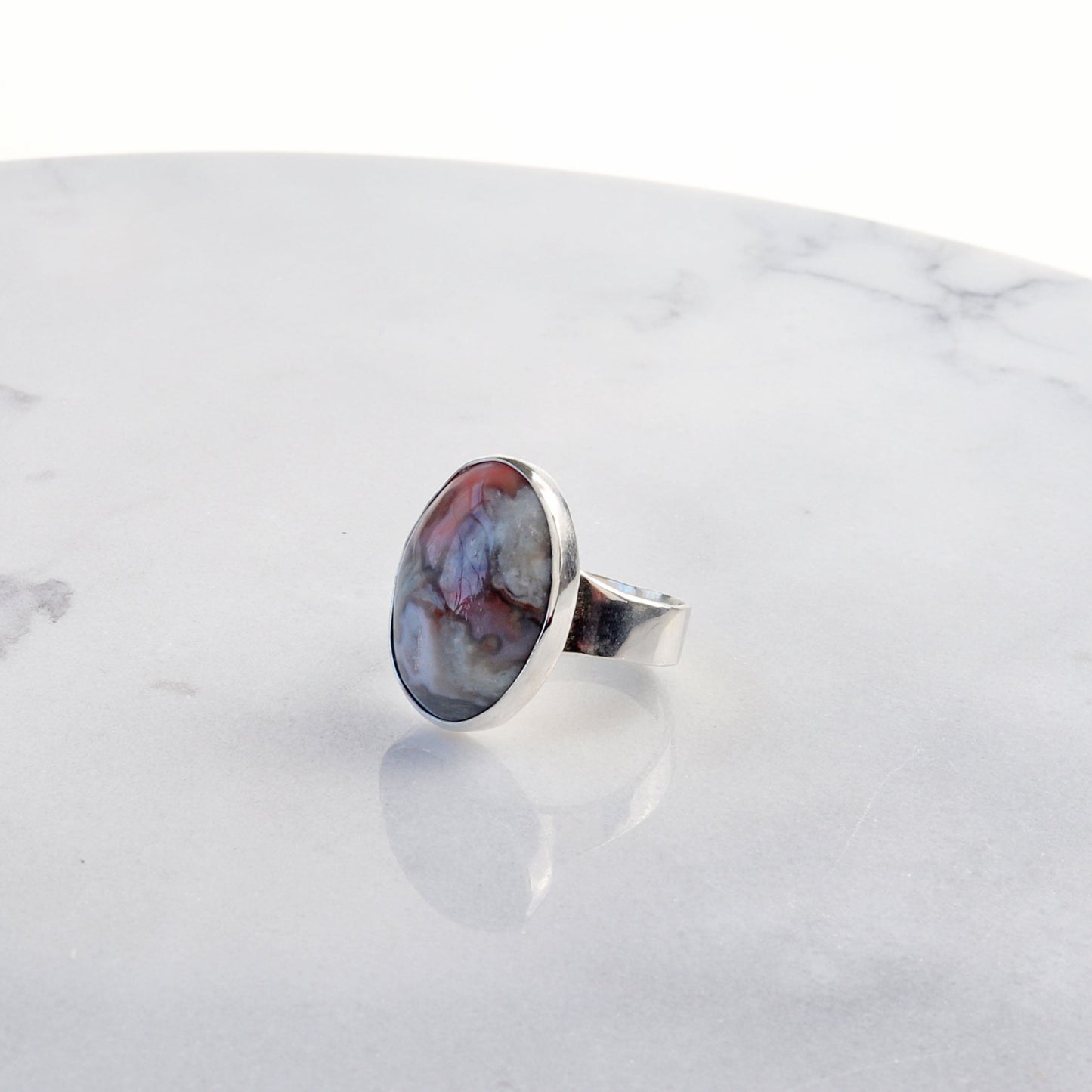 A Crazy Lace Agate Natural Cabochon Gemstone Ring. Handcrafted to reveal the unique swirling patterns and vibrant hues of pink and gray and a unique Crystal feature within the stone. Encased in sterling 935 Argentium silver with a fine silver bezel (.999).