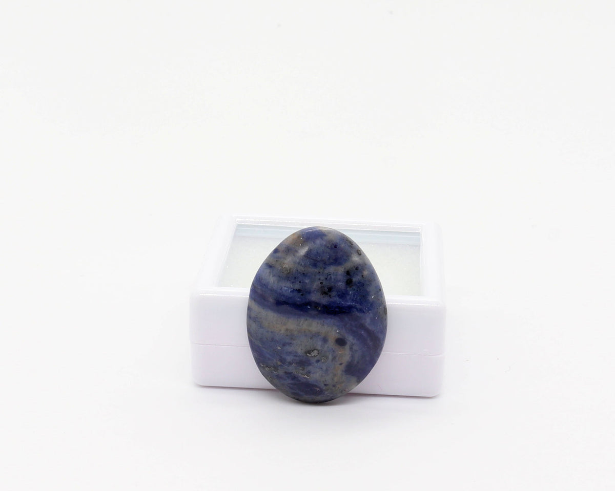 A hand-cut and polished natural Foidolite cabochon with layered deep blue, slate blue and gray natural mineral patterning, displayed at side angle against a white gem box, photographed on a clean white background.