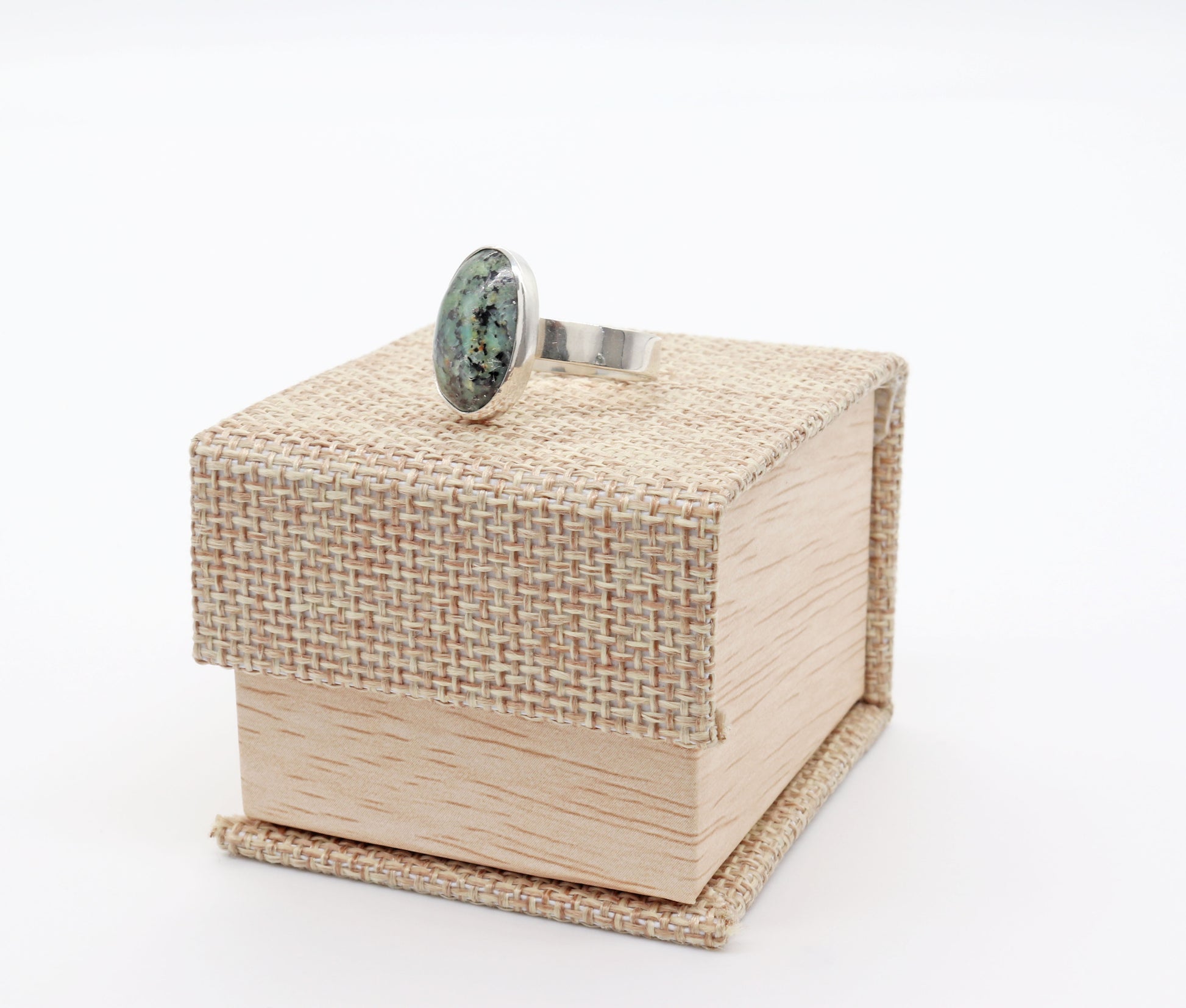 A Fraser River Turquoise ring shown on Cabana Burlap and Wood Patterned Ring Gift Box included with purchase.  The ring features a handcut and polished gemstone with stunning blue-green hues and bold black mottled matrix, echoing the rugged landscape of the Fraser River region in British Columbia