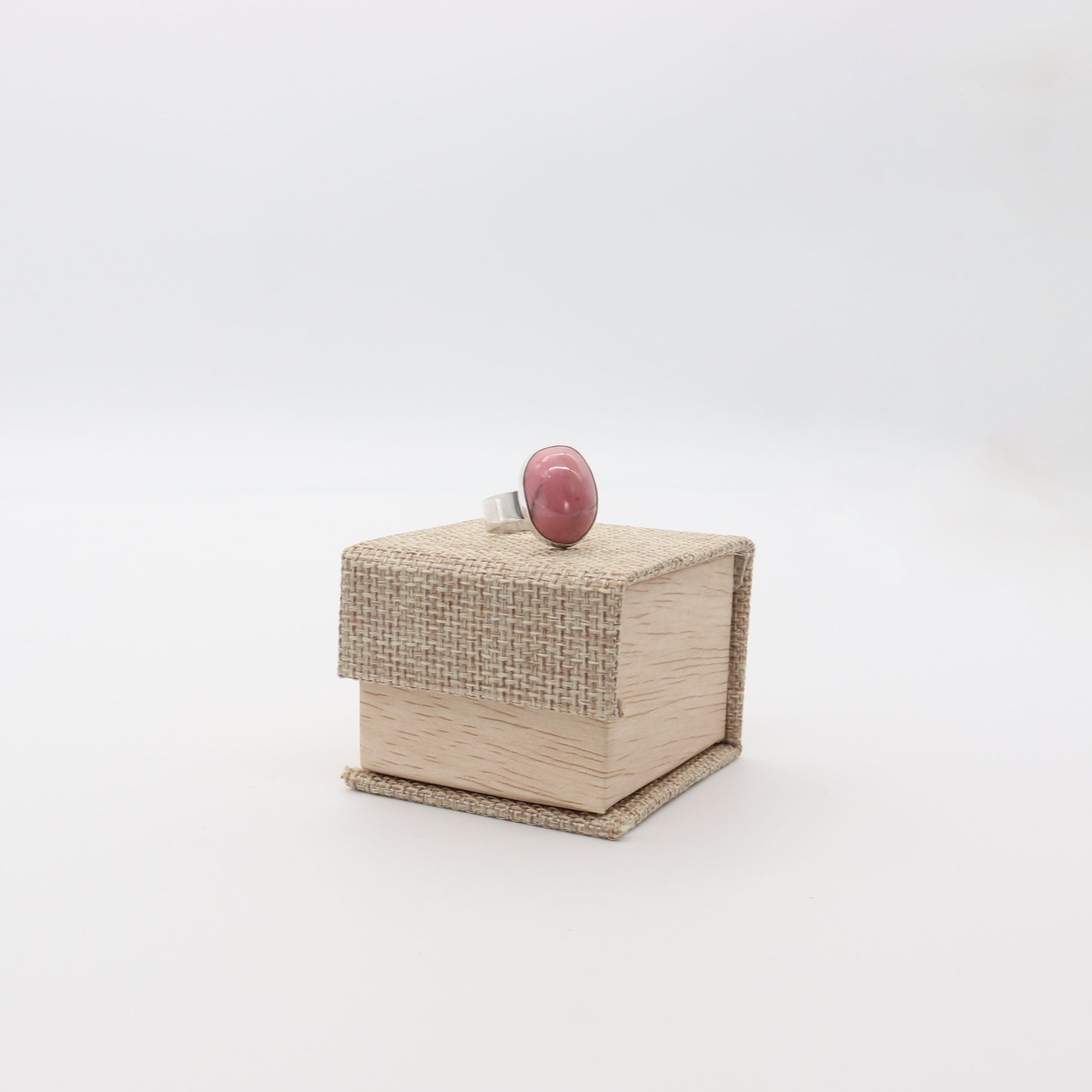 A natural pink rose Rhodonite gemstone Ring, handcut and polished into a smooth cabochon and encased in sterling 935 Argentium silver with a fine silver bezel (.999) shown on a CABANA Burlap and Wood Patterned Ring Gift Box.