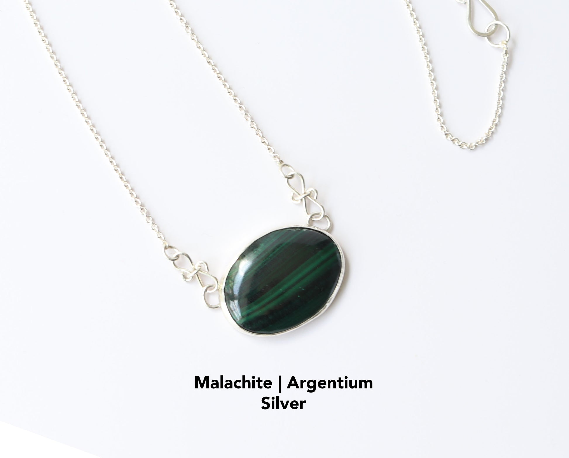 A unique Malachite gemstone pendant hand-cut and polished stone and set in solid sterling 935 Argentium® Silver. Designed with a custom chain and S hook for easy wearability. Shown against a white marble background.