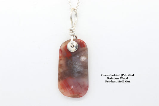 A one-of-a-kind pendant features a stunning tag-shaped cabochon of Petrified Rainbow Wood, hand-cut and polished to reveal warm hues of red, mauve, white, and translucent veining with Argentium Silver bail and chain.