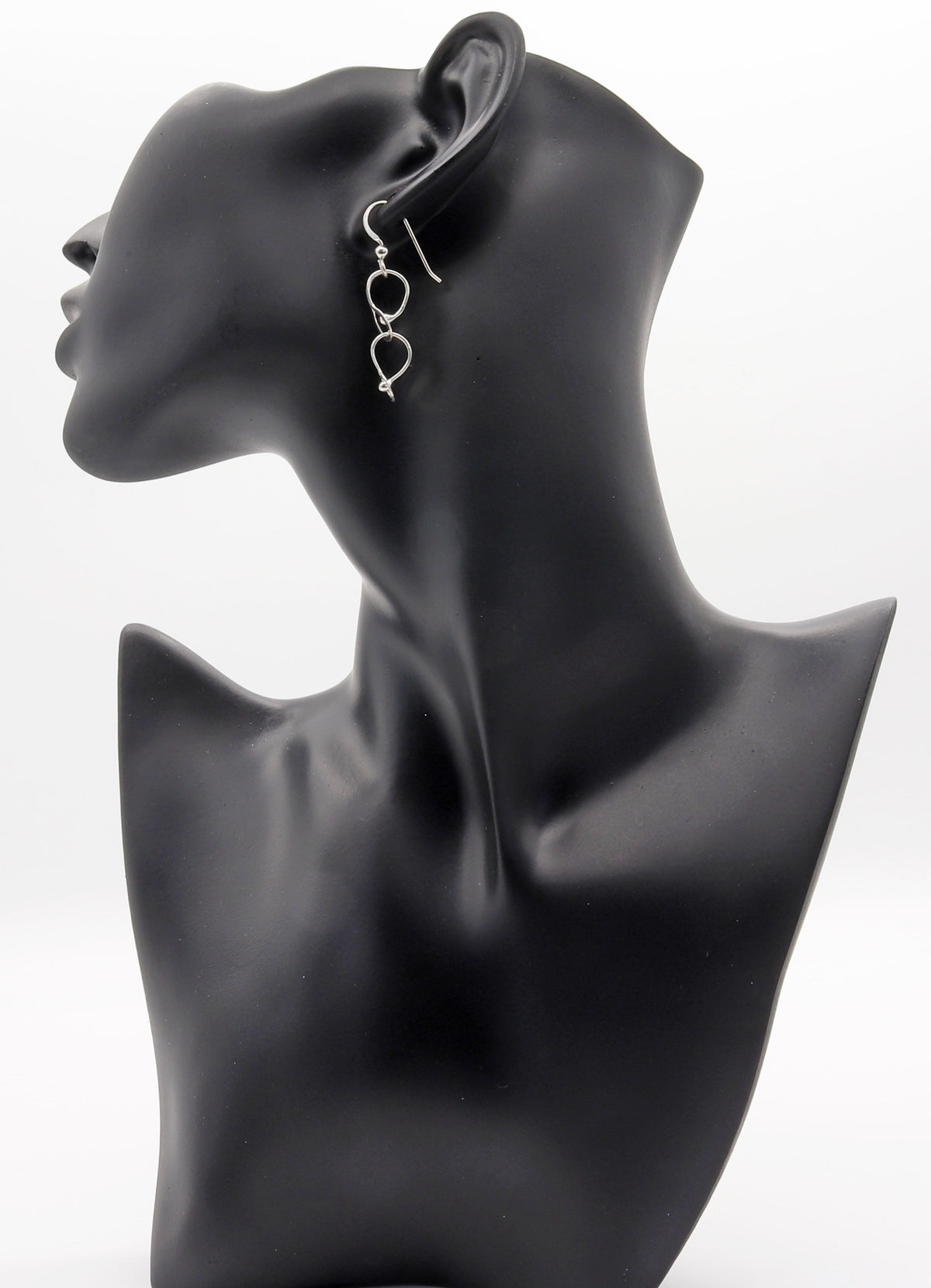 A pair of silver dangling hoop earrings with small heart charms, displayed on a black mannequin head against a white studio background.