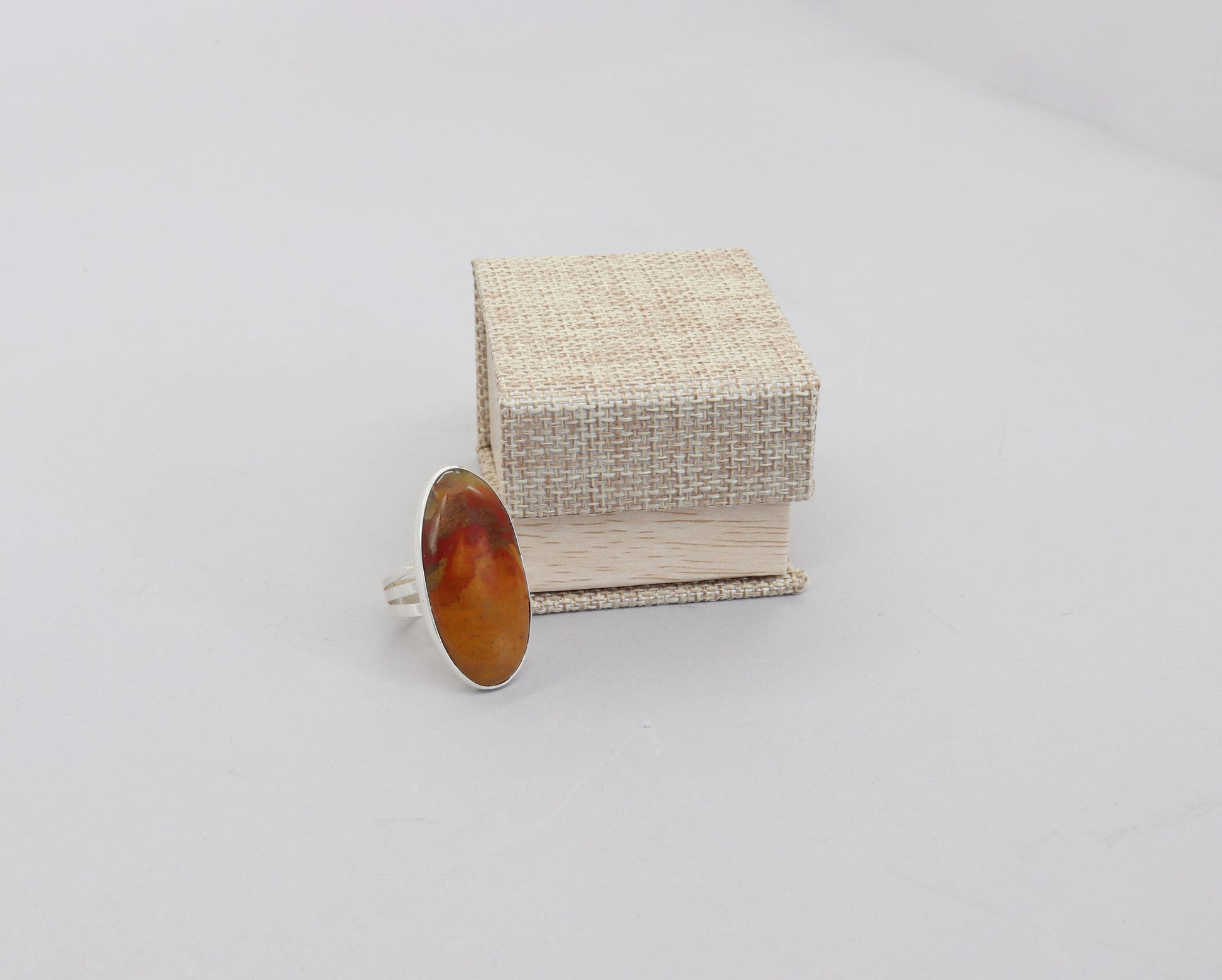 A sunrise sonoran gemstone ring set in Argentium silver.  Handcut and polished to reveal the stunning hues of, brown, orange, and red gemstone. The ring is sitting on a white surface next to a Burlap and Wood Patterned Ring Gift Box, ready to gift.