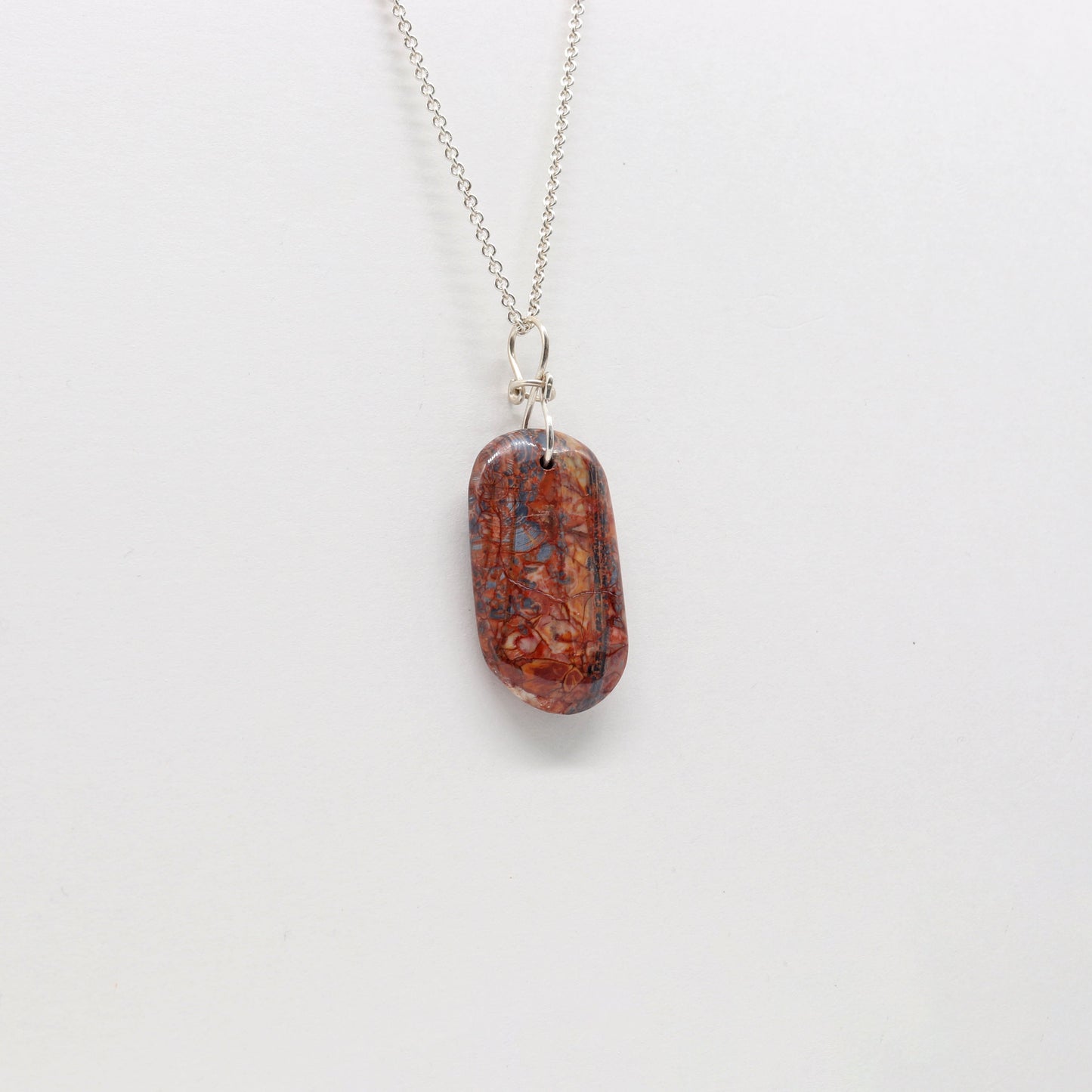A Stony Creek Jasper gemstone pendant. Handcut and polished on both sides to showcase the unique red and blue patterns of the stone.  The pendant is designed with an original infinity bow shaped wire bail made from Argentium® 935 Silver that is tarnish resistance and has a remarkable shine.  To complete the look a complementary 935 Argentium® silver chain is included.