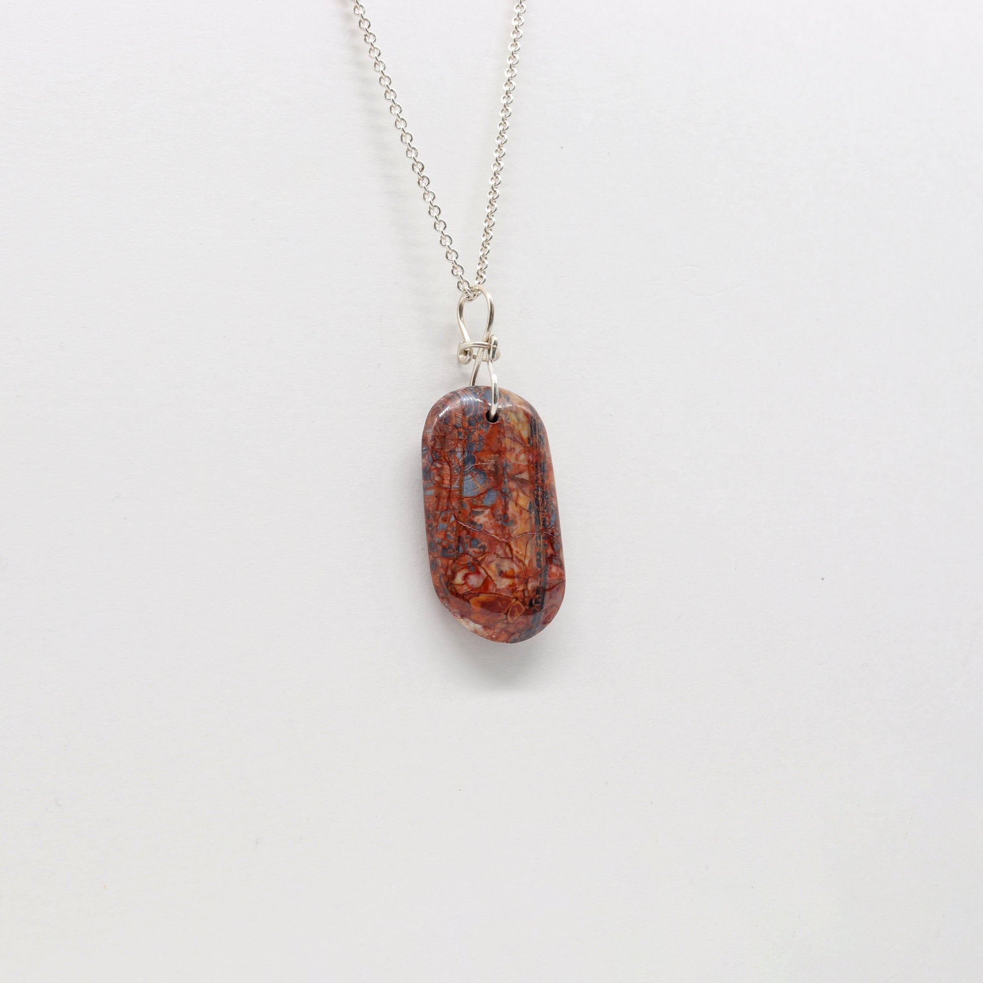 A Stony Creek Jasper gemstone pendant. Handcut and polished on both sides to showcase the unique red and blue patterns of the stone.  The pendant is designed with an original infinity bow shaped wire bail made from Argentium® 935 Silver that is tarnish resistance and has a remarkable shine.  To complete the look a complementary 935 Argentium® silver chain is included.
