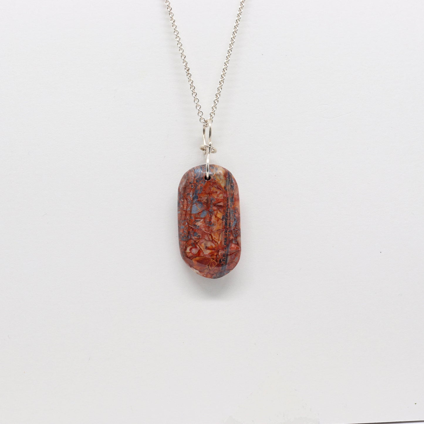 A Stony Creek Jasper gemstone pendant. Handcut and polished on both sides to showcase the unique red and blue patterns of the stone.  The pendant is designed with an original infinity bow shaped wire bail made from Argentium® 935 Silver that is tarnish resistance and has a remarkable shine.  To complete the look a complementary 935 Argentium® silver chain is included.