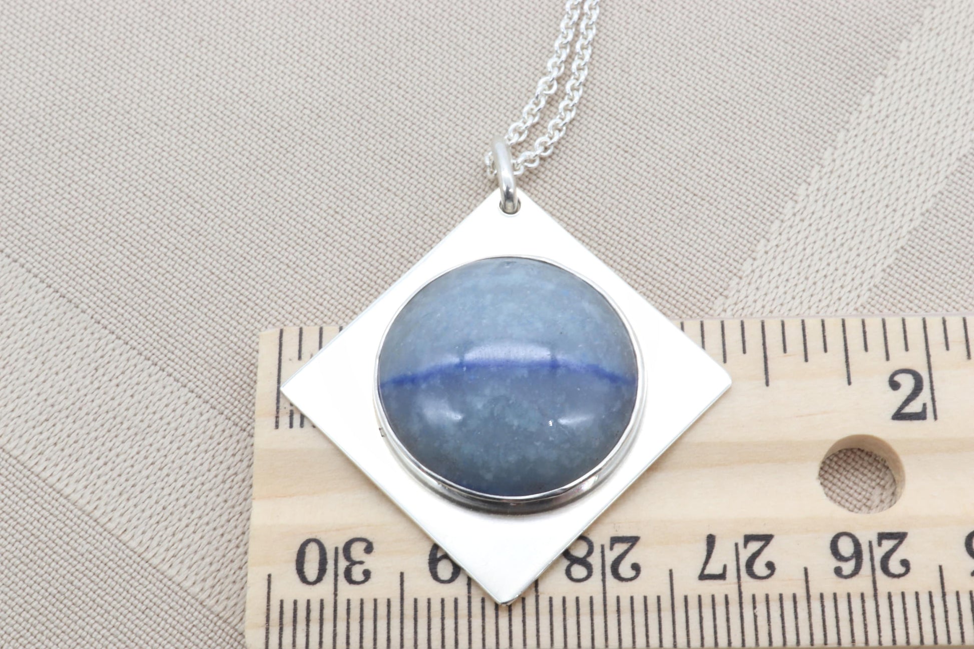A 1.375 inch Celestial Blue Aventurine pendant with Saturn-like inclusions.   Shown placed on a ruler to demonstrate the size perspective on a background of beige linen with a chain necklace attached to the pendant.