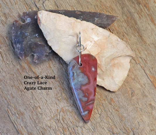 A one-of-a-kind Crazy Lace Agate arrowhead shaped focal charm, polished on both sides to reveal swirling patterns in mauve, deep reds, and grays. Shown against driftwood an real arrowheads as backdrop props.