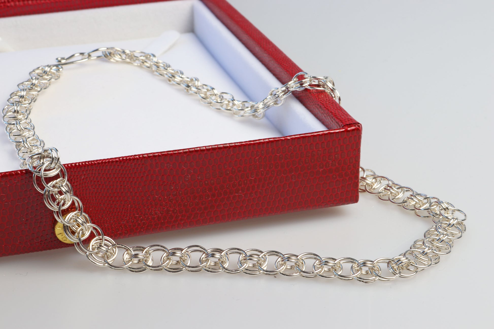 Red faux leather gift box open against white background with helm weave chain draped over the corner of the box.  Box is included in purchase of helm weave chain.
