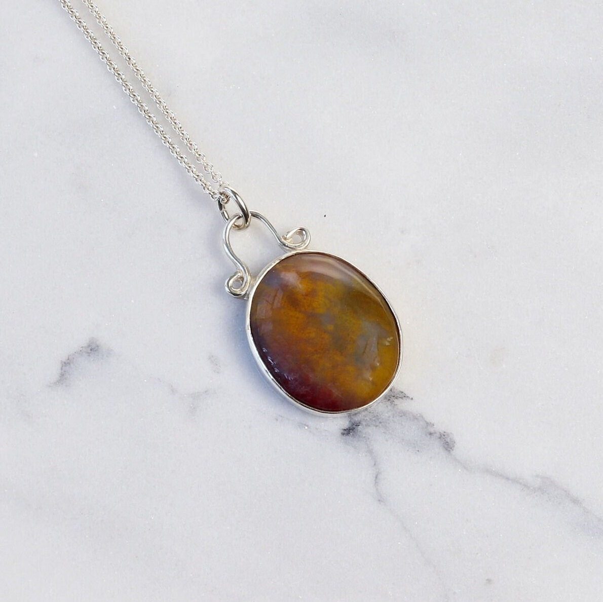 A Petrified Rainbow Wood pendant handmade from a raw Arizona Petrified Wood rock, carefully cut and polished to reveal gold, gray and deep red hues showcased in a vintage style silver setting. Hanging from a unique and elegant sterling 935 Argentium Silver bail and setting shown against white marble background.