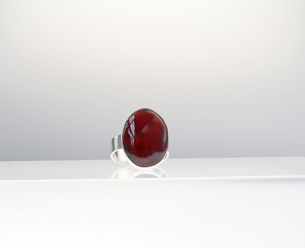 A front view of Handcrafted Petrified Rainbow Wood ring with oval red and dark brown gemstone set in polished Argentium 935 silver, displayed on a white block against a light gray background.