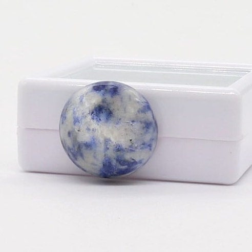 Close-up view of a polished, round-shaped blue sodalite cabochon gemstone. The stone exhibits a captivating blend of blue and white hues, creating a marbled effect.  The gemstone is displayed in a simple, white gem box display case with glass lid, for storage for gifting. The overall impression is one of elegance and natural beauty, highlighting the unique characteristics of this cabochon. This image showcases a high-quality, natural gemstone, ideal for jewelry making or collectors.