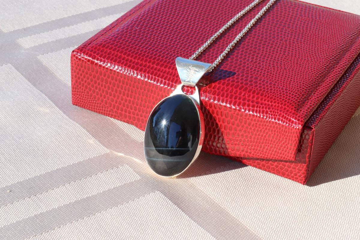 An oval natural Blue Tiger’s Eye pendant in a polished Argentium 935 silver bezel, suspended from a silver chain resting on a textured red box over a beige striped fabric background.