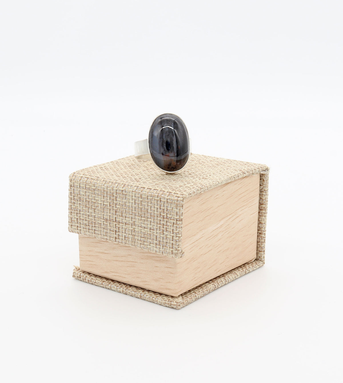 Handcrafted Petrified Rainbow Wood ring with polished blue-black oval gemstone set in solid Argentium 935 silver with fine silver bezel displayed on a light wood and woven fabric jewelry box against a white background.