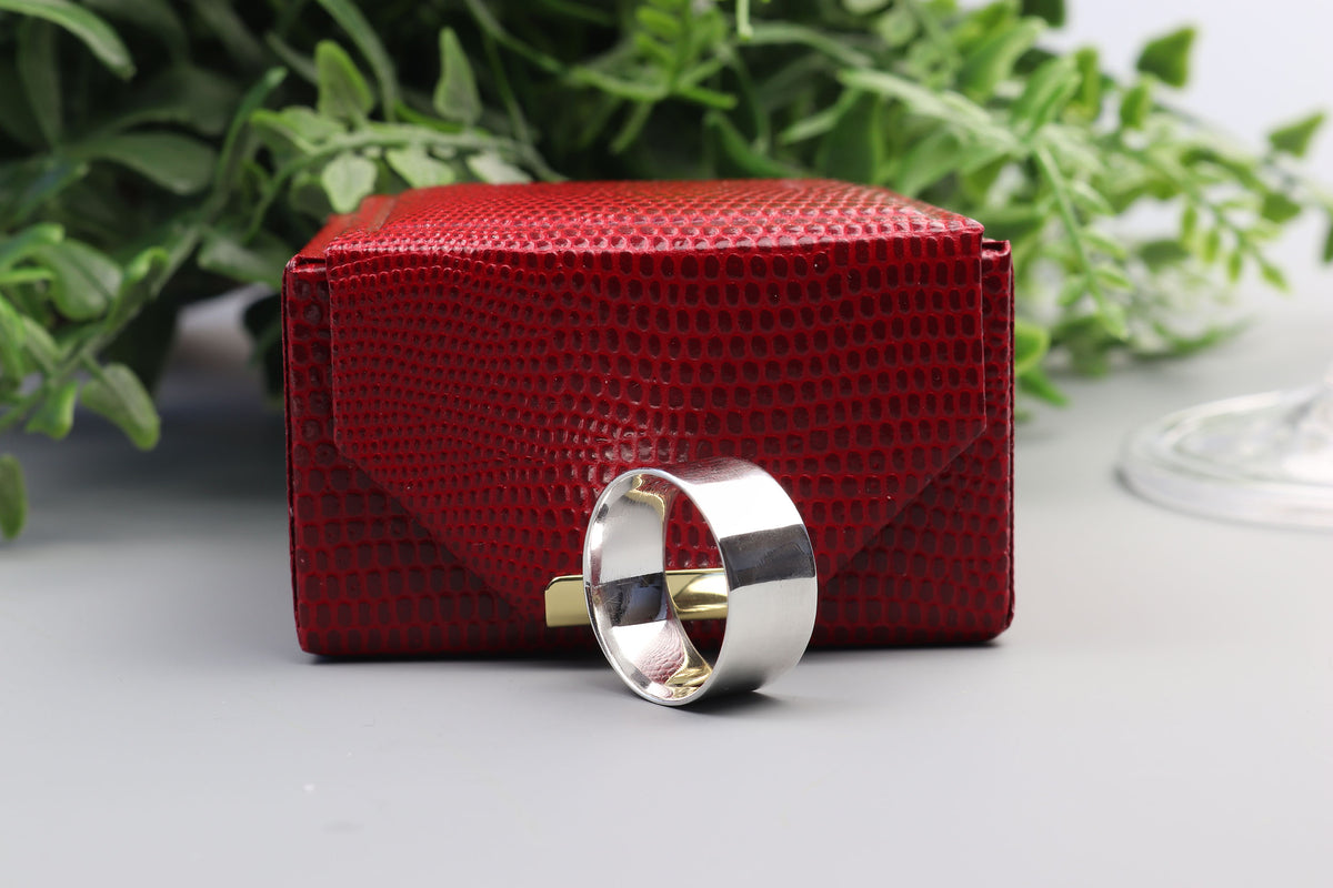 Solid Argentium 935 silver wide band ring with polished anticlastic contour, displayed in front of a textured red gift box with green foliage in the background.