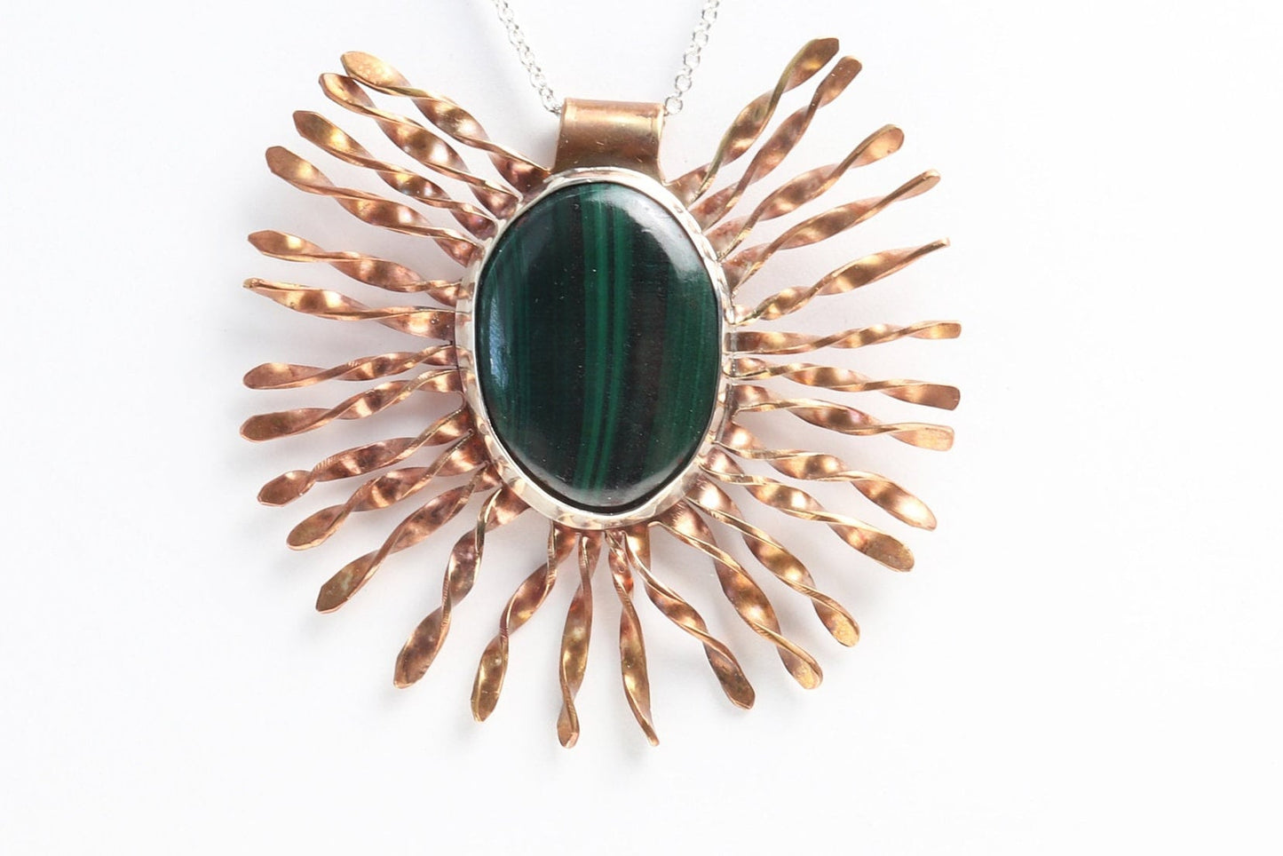 A unique Malachite Gemstone Pendant handcrafted in our studio. The Malachite gemstone has been handcut and polished into a cabochon and encased in a sterling 935 Argentium silver setting. A unique starburst pattern in Jewelers Brass with a captivating copper patina, elegantly encircles the polished malachite stone.