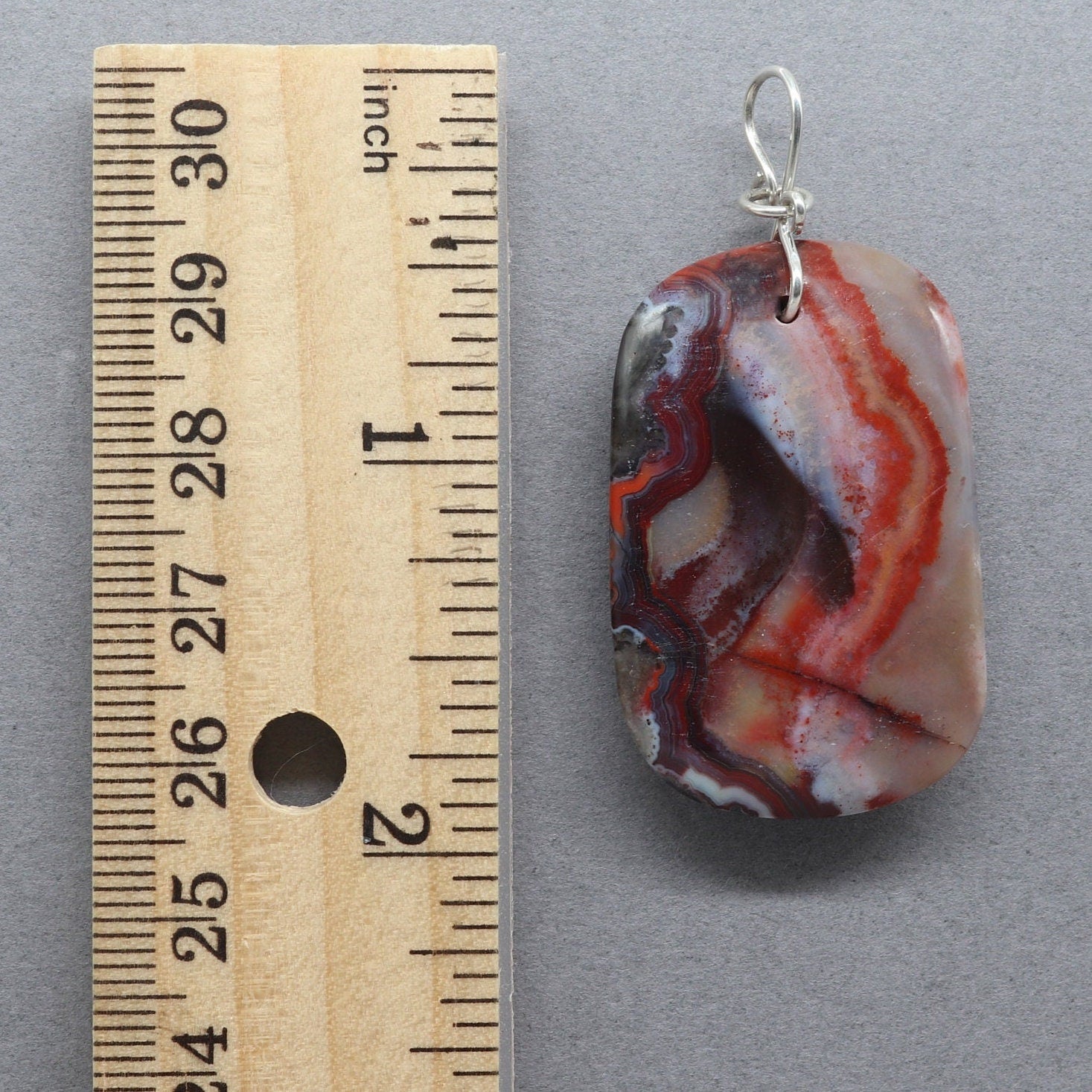 A 1.95 inch Red Mexico Agate hand-cut and freeform polished to bring out the mauve gray and deep red patterns and translucent colors. Crafted with an original infinity bow wire bail in Argentium 935 silver known for its higher silver content and its ability to resist tarnishing and keep on shining.