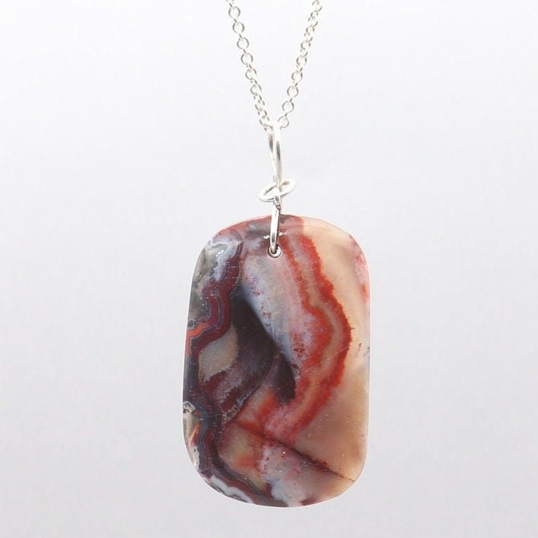 A one-of-a-kind Red Mexico Agate hand-cut and freeform polished to bring out the mauve gray and deep red patterns and translucent colors. Crafted with an original infinity bow wire bail in Argentium 935 silver known for its higher silver content and its ability to resist tarnishing and keep on shining.