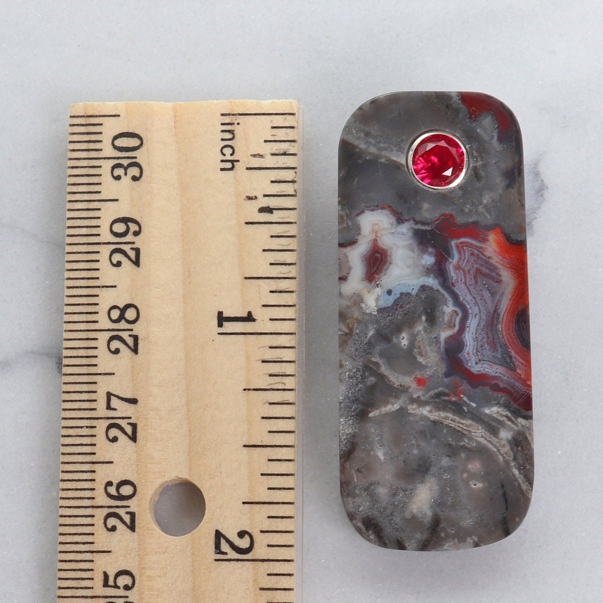 A 2 inch tag shaped Mexico Agate, handcut and freeform polished with a 6mm lab ruby inlay. The pendant has been fashioned with a bail in premium sterling 935 Argentium® Silver, renowned for its resistance to tarnishing and its brilliant luster. To complete the look is a complementary handcrafted leather cord necklace with Nu Gold Brass S hook clasp.