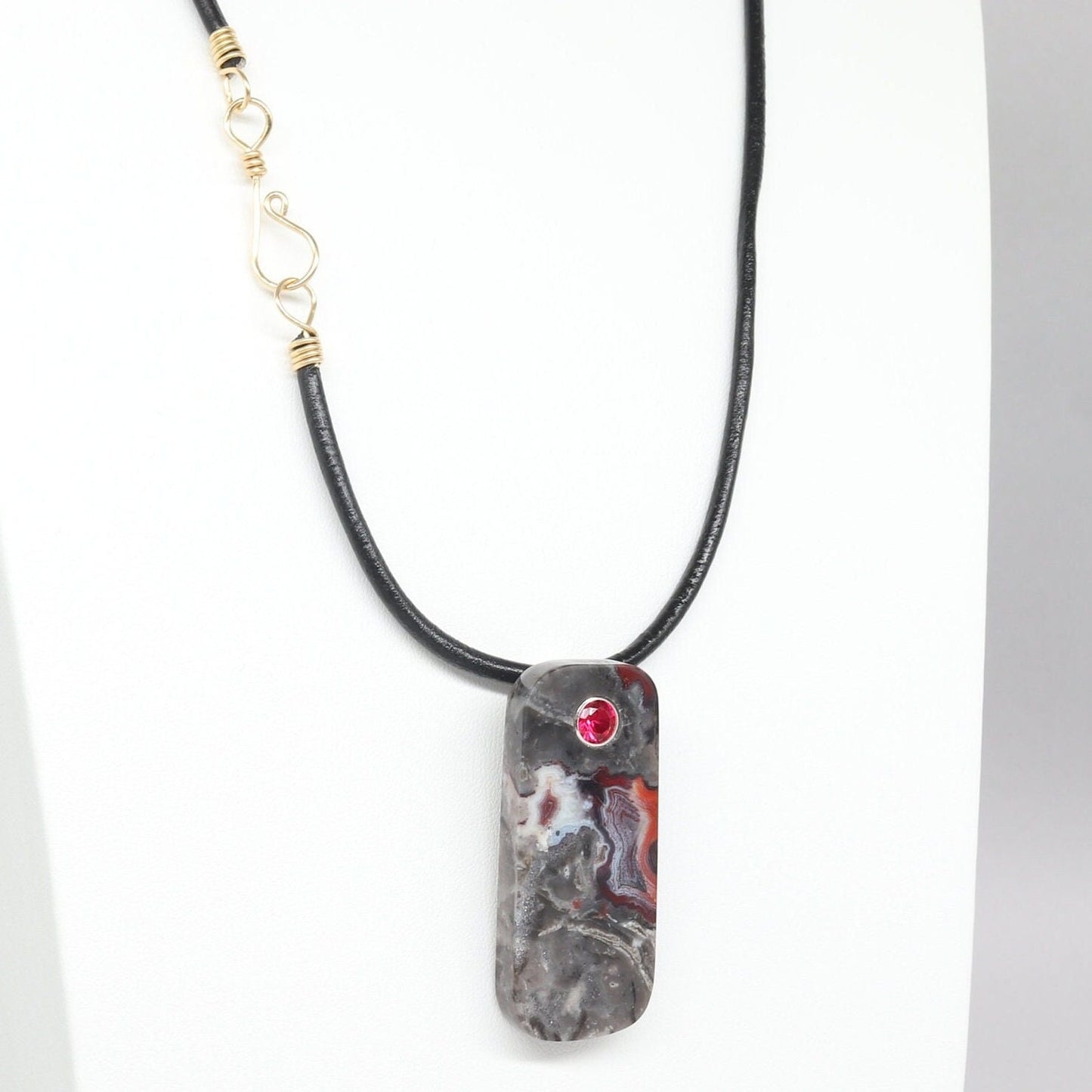 A 2 inch tag shaped Mexico Agate, handcut and freeform polished with a 6mm lab ruby inlay. The pendant has been fashioned with a bail in premium sterling 935 Argentium® Silver, renowned for its resistance to tarnishing and its brilliant luster. To complete the look is a complementary handcrafted leather cord necklace with Nu Gold Brass S hook clasp.