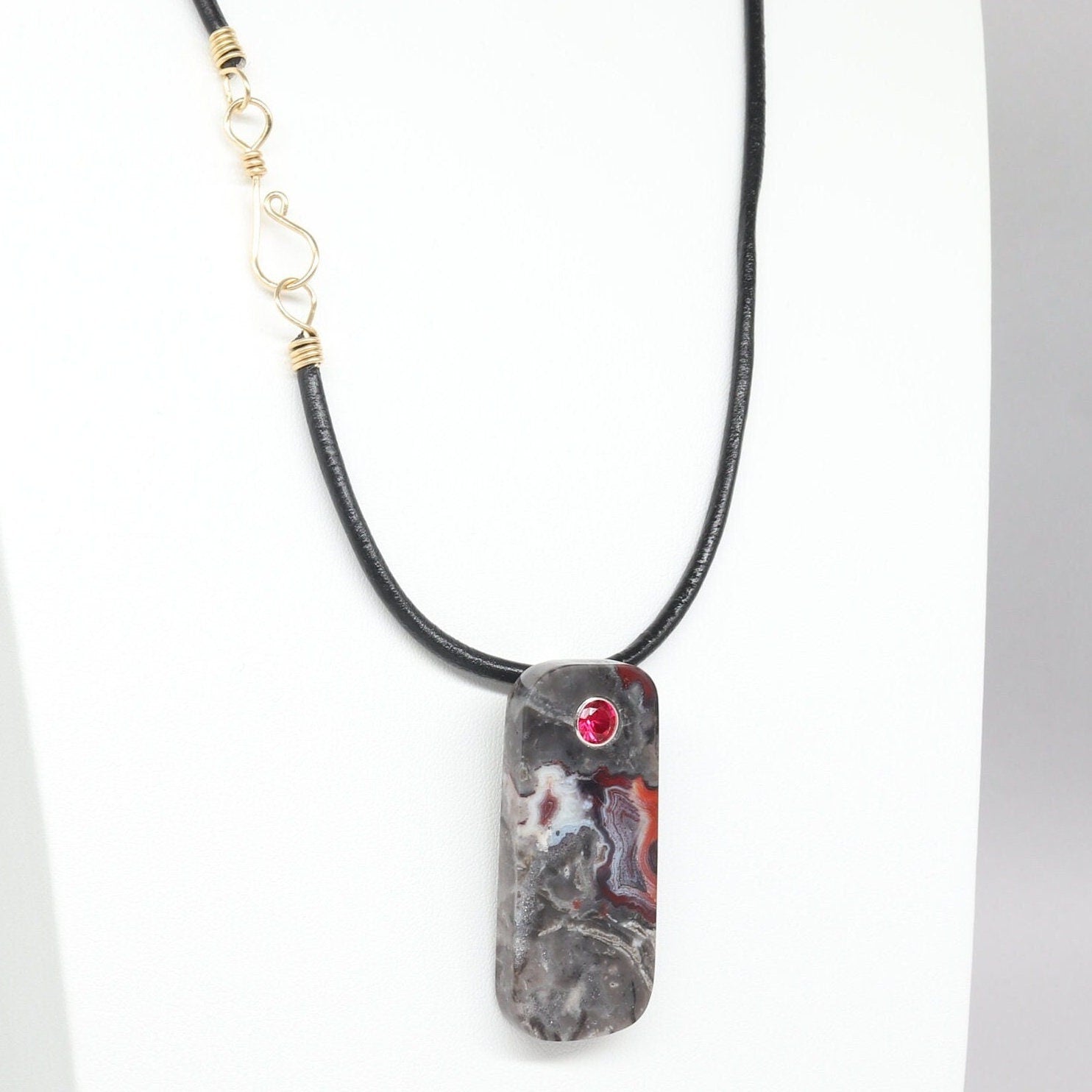 A 2 inch tag shaped Mexico Agate, handcut and freeform polished with a 6mm lab ruby inlay. The pendant has been fashioned with a bail in premium sterling 935 Argentium® Silver, renowned for its resistance to tarnishing and its brilliant luster. To complete the look is a complementary handcrafted leather cord necklace with Nu Gold Brass S hook clasp.
