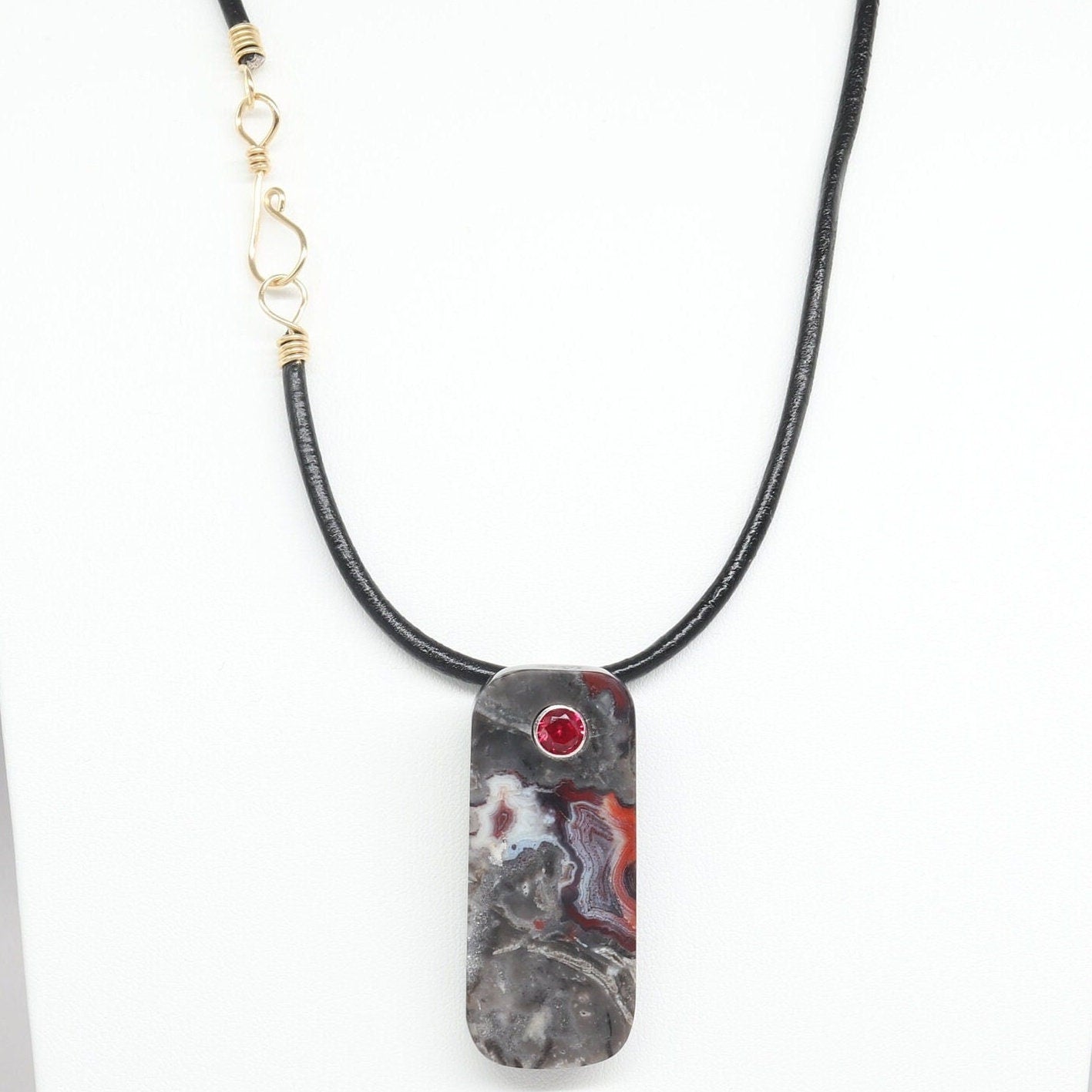 A tag shaped Mexico Agate, handcut and freeform polished with a 6mm lab ruby inlay. The pendant has been fashioned with a bail in premium sterling 935 Argentium® Silver, renowned for its resistance to tarnishing and its brilliant luster. To complete the look is a complementary handcrafted leather cord necklace with Nu Gold Brass S hook clasp.