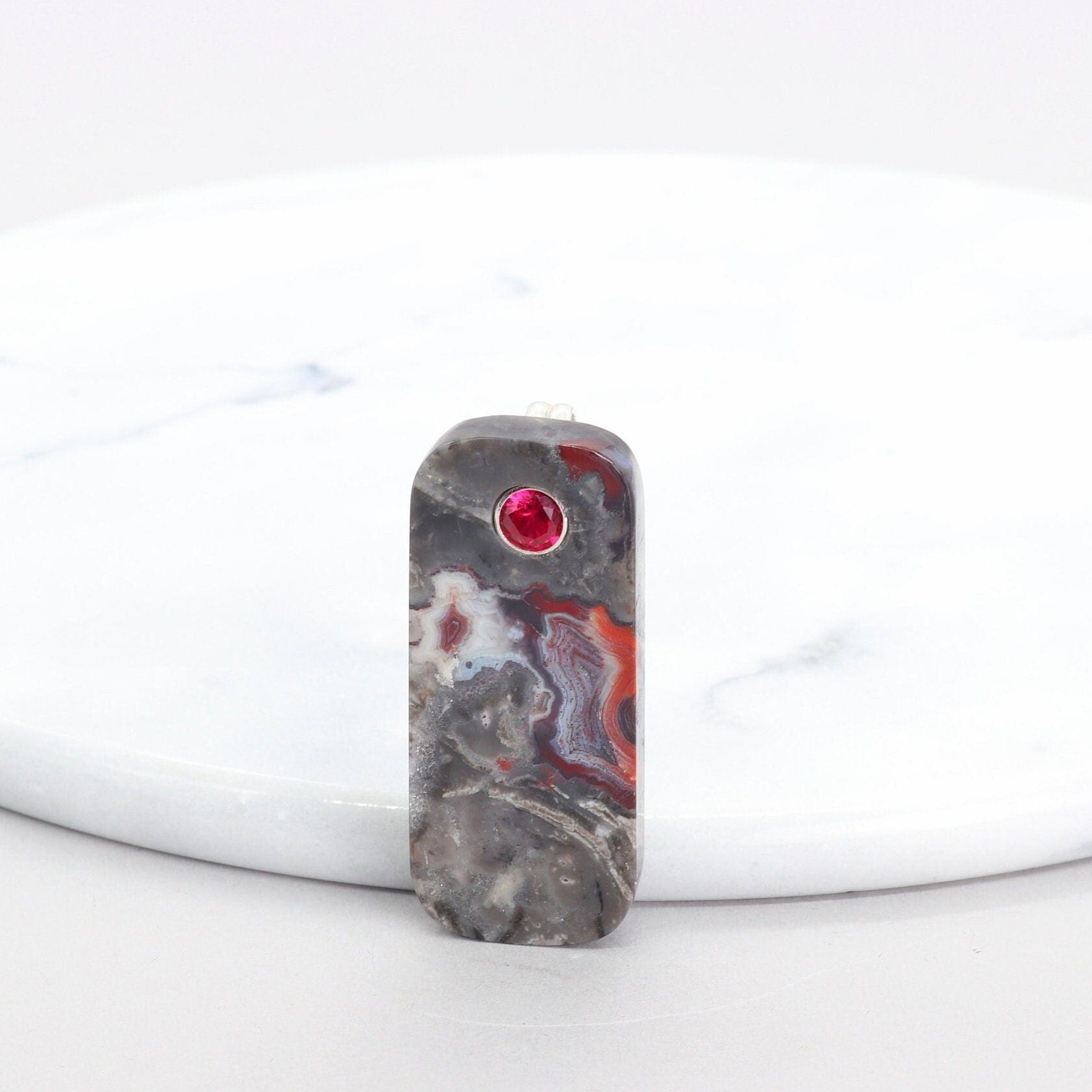 A tag shaped Mexico Agate, handcut and freeform polished with a 6mm lab ruby inlay. The pendant has been fashioned with a bail in premium sterling 935 Argentium® Silver, renowned for its resistance to tarnishing and its brilliant luster. To complete the look is a complementary handcrafted leather cord necklace with Nu Gold Brass S hook clasp.