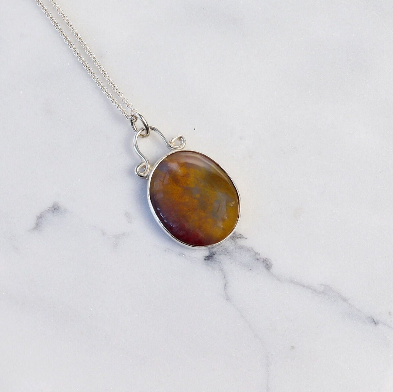 A Petrified Rainbow Wood stone found in Arizona's Petrified Forest, carefully polished by hand in our studio to reveal gold, gray and deep red hues showcased in a vintage style setting. Hanging from a unique and elegant sterling 935 Argentium Silver bail and setting, this piece is sure to capture your attention and become a cherished part of your jewelry collection.