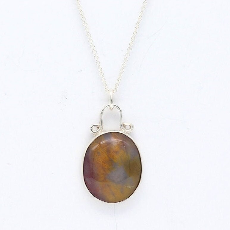 A Petrified Rainbow Wood stone found in Arizona's Petrified Forest, carefully polished by hand in our studio to reveal its stunning charm and vintage appearance. Hanging from a unique and elegant sterling 935 Argentium® Silver bail and setting, this piece is sure to capture your attention and become a cherished part of your jewelry collection.