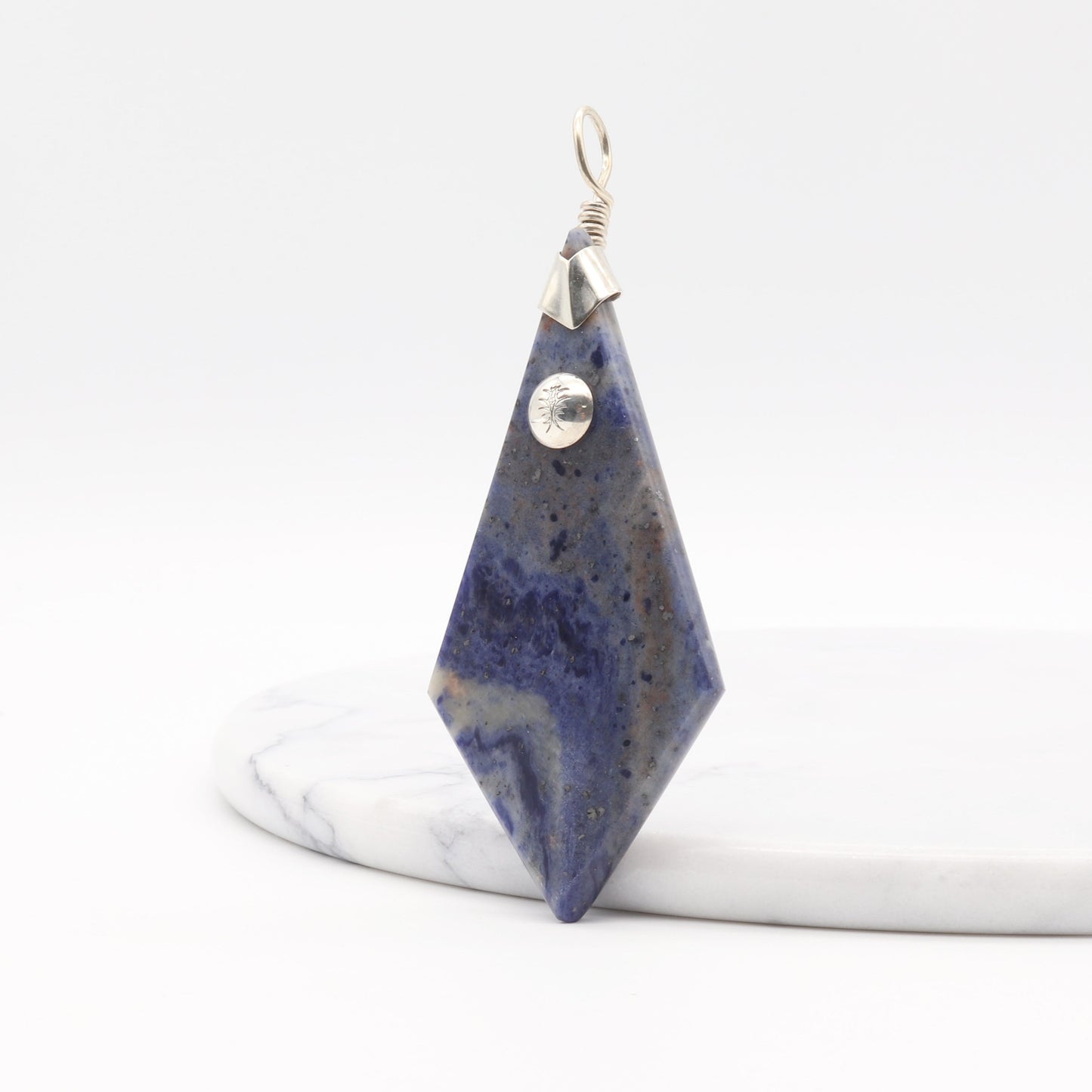 Side view of Blue Foidolite pendant, with a handcrafted Argentium® 935 Silver bail—against a white marble tray on white background.