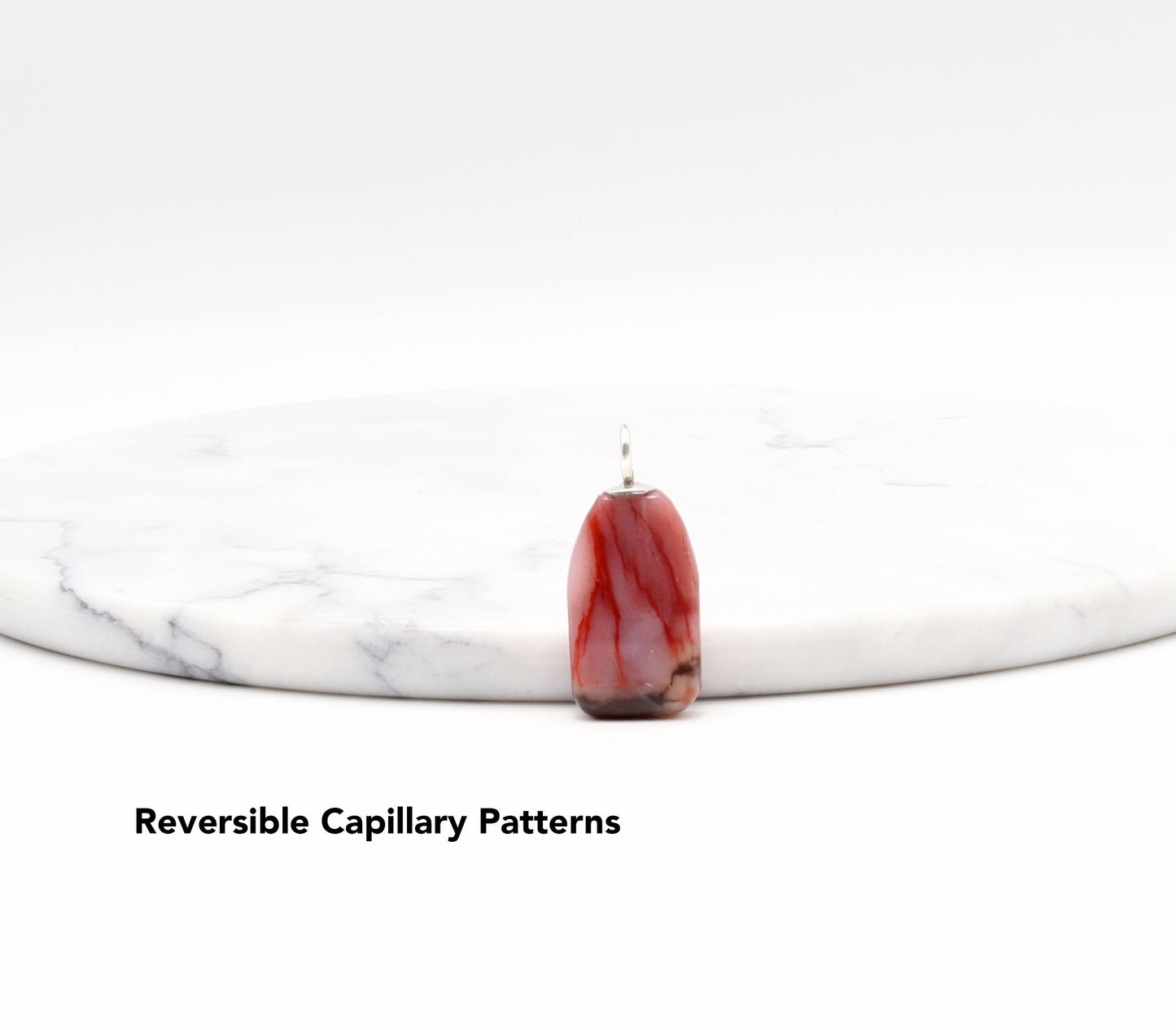 Red and black petrified rainbow wood pendant on a white marble surface with 'Reversible Capillary Patterns' text.