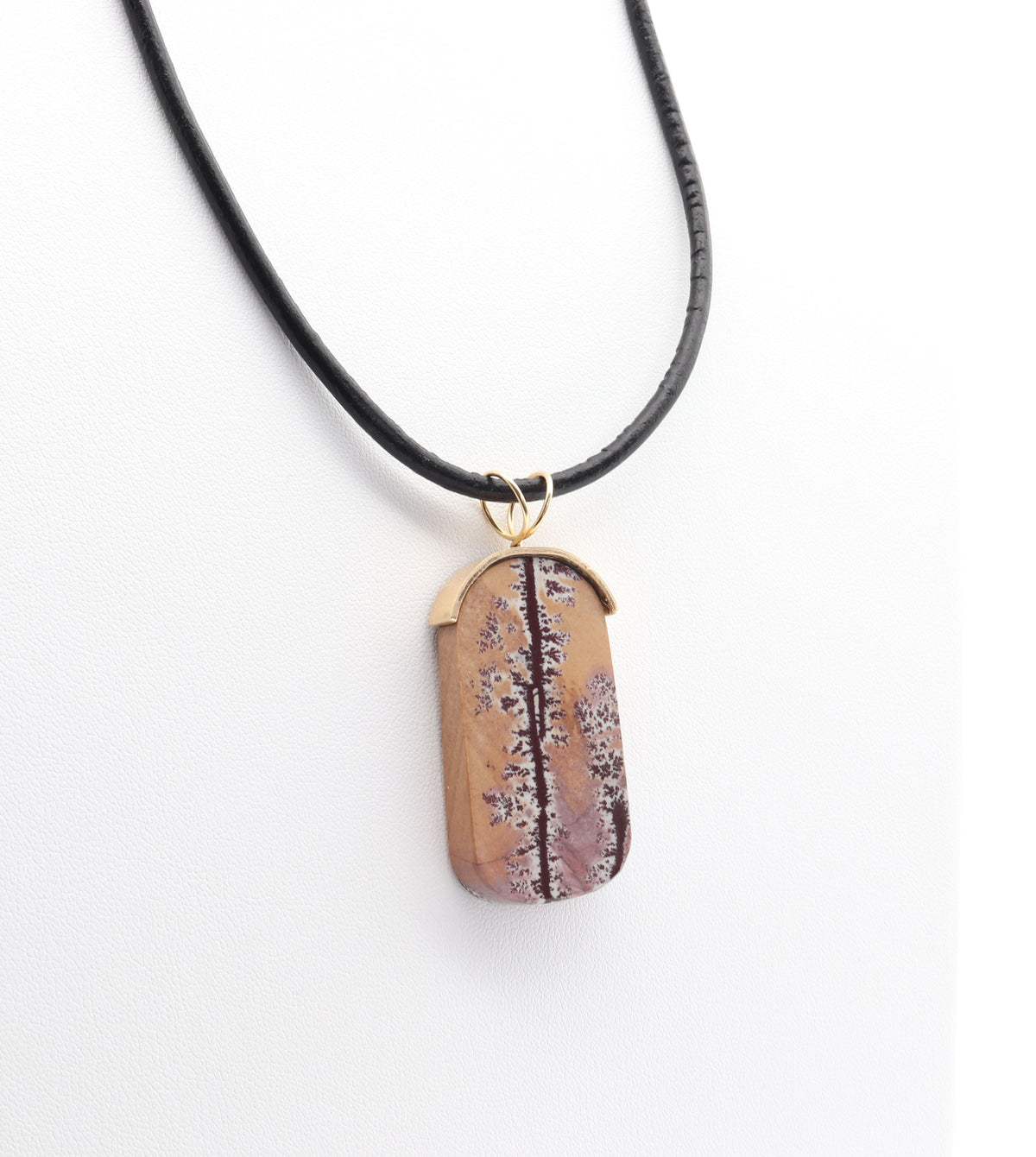 A one-of-a-kind tag-shaped Sonoran Dendritic Rhyolite pendant with a black leather cord, featuring a tan and brown wood-grain patterned pendant with purple and white floral details, and a gold-tone metal cap. Shown on white jewelry form against white background.