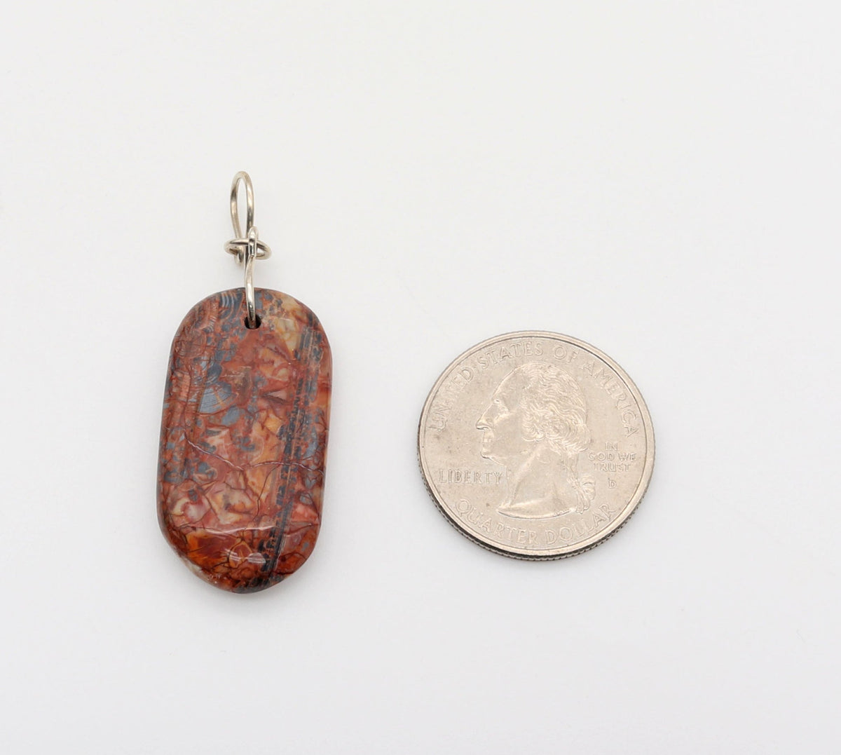 A small oval Stony Creek pendant with a reddish-brown marbled pattern and a silver bail, shown next to a US quarter coin for scale on a plain white background.