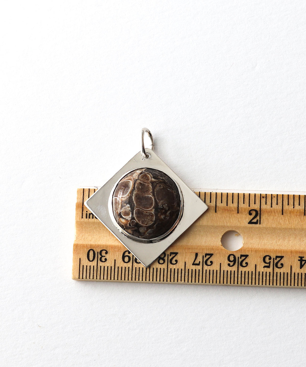A silver Turritella stone pendant with a square bezel containing a round brown and black patterned stone, shown next to a wooden ruler on a white background.