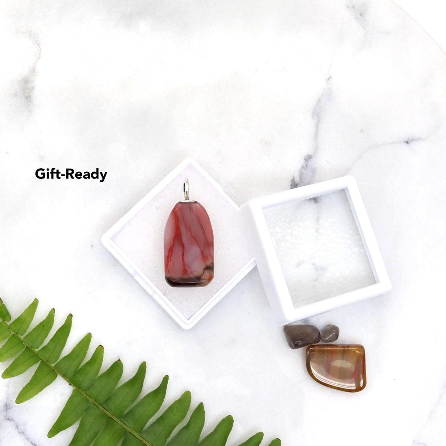 Hand-cut and polished on both sides in our Missouri studio, this tag-shaped gemstone reveals stunning red, pink, black and translucent patterns, with intricate capillary-like designs resembling nature’s delicate vein structures. Shown in white gem box with glass lid ready for gifting or storage.