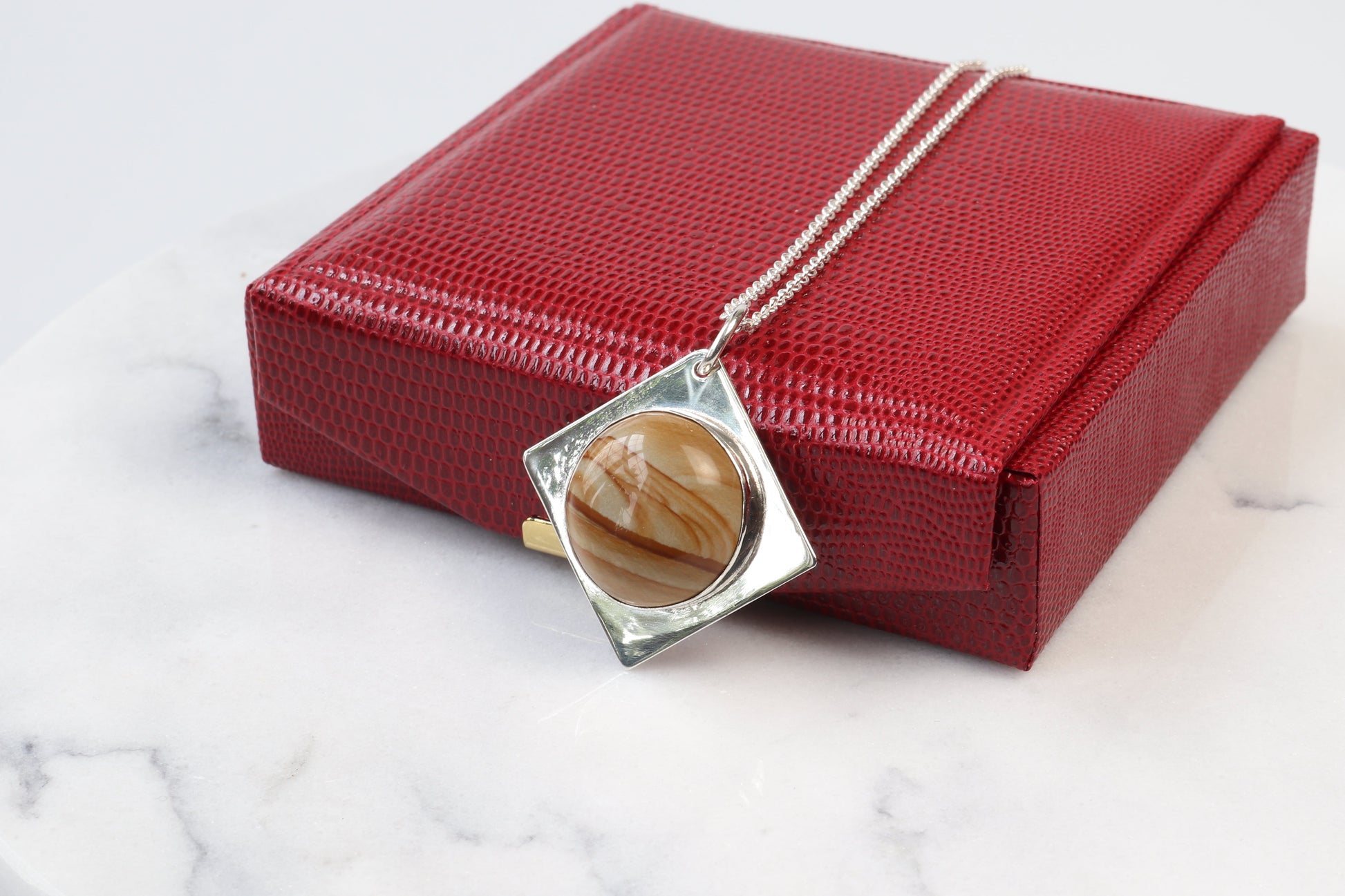 A 1.625 inch hand-cut and polished one-of-a-kind Picture Jasper cabochon with a beautiful landscape pattern and color bands set in a unique four-point diamond-shaped of sterling Argentium® 935 Silver paired with a complementary sterling Argentium 935 chain.  Ready to gift in a red faux leather gift box.