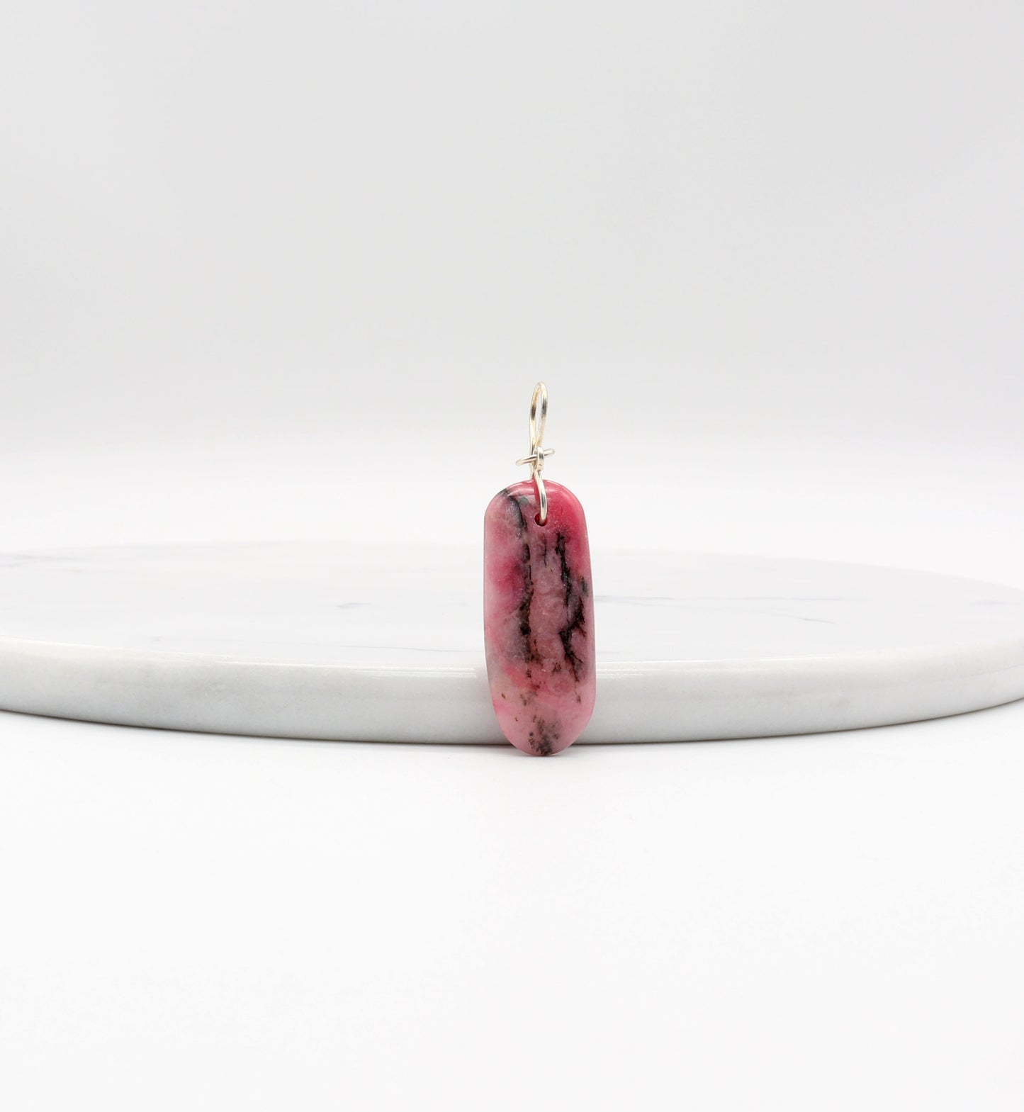 A pink rhodonite gemstone with black inclusions designed with a silver bail. The stone is a long oval shape with a smooth surface. Shown against a white marble slab.