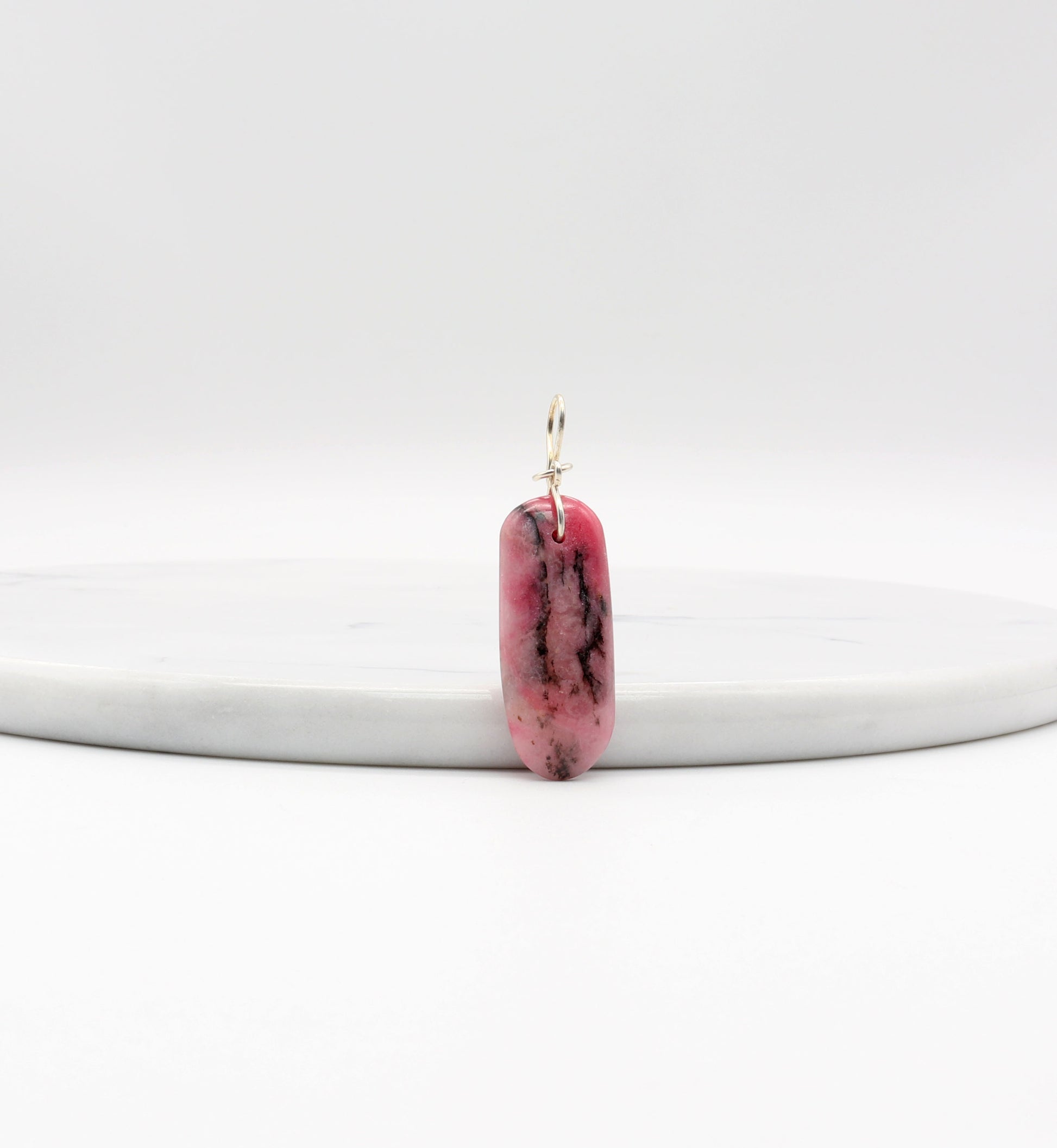 A pink rhodonite gemstone with black inclusions designed with a silver bail. The stone is a long oval shape with a smooth surface.  Shown against a white marble slab.