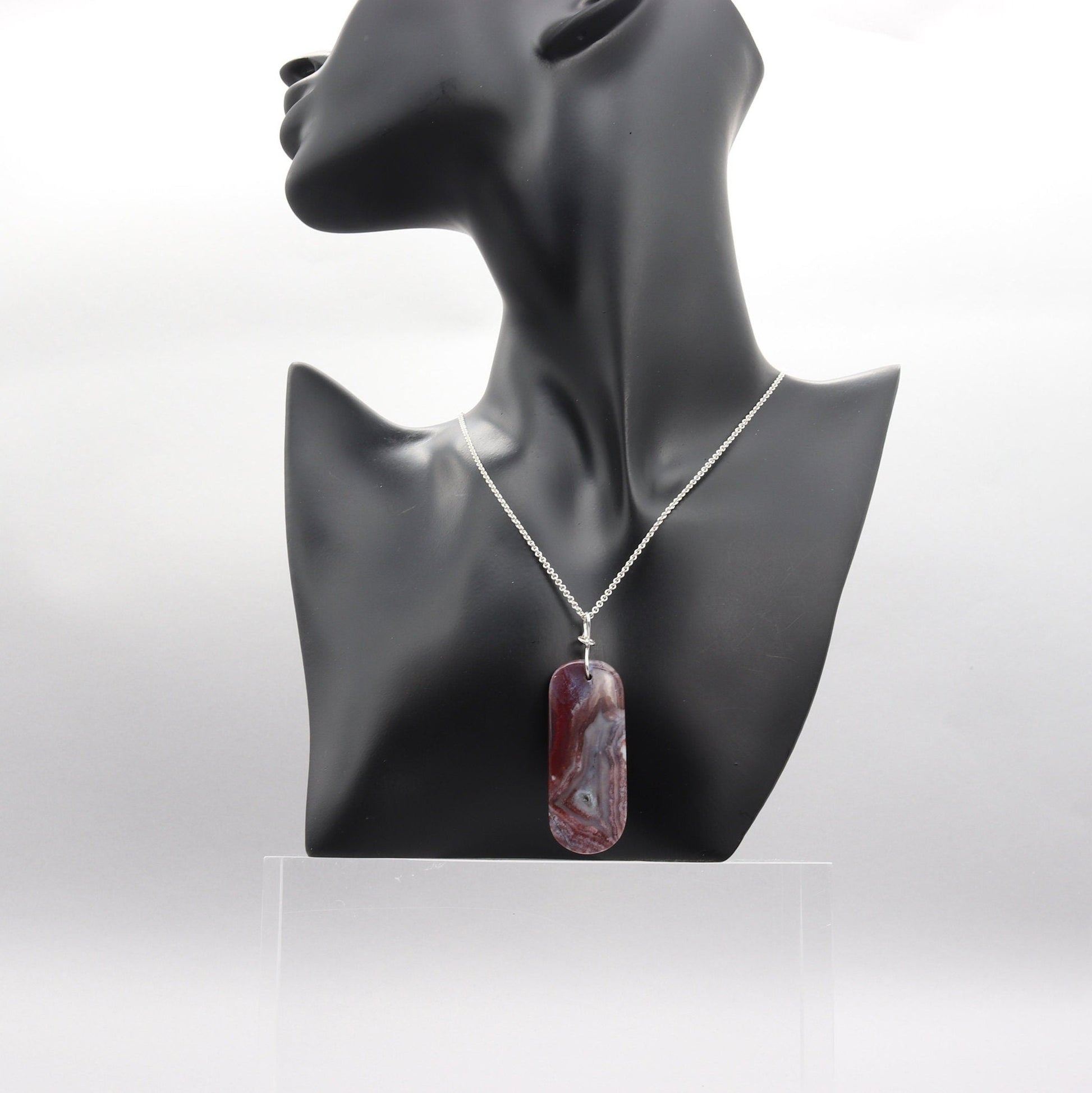 A one-of-a-kind Red Mexico Agate pendant with hues of mauve gray and deep lavender. Hand-cut and expertly freeform polished to accentuate the charming small crystal feature and stunning translucent patterns. Shown with Argentium necklace on black resin mannequin against white background.