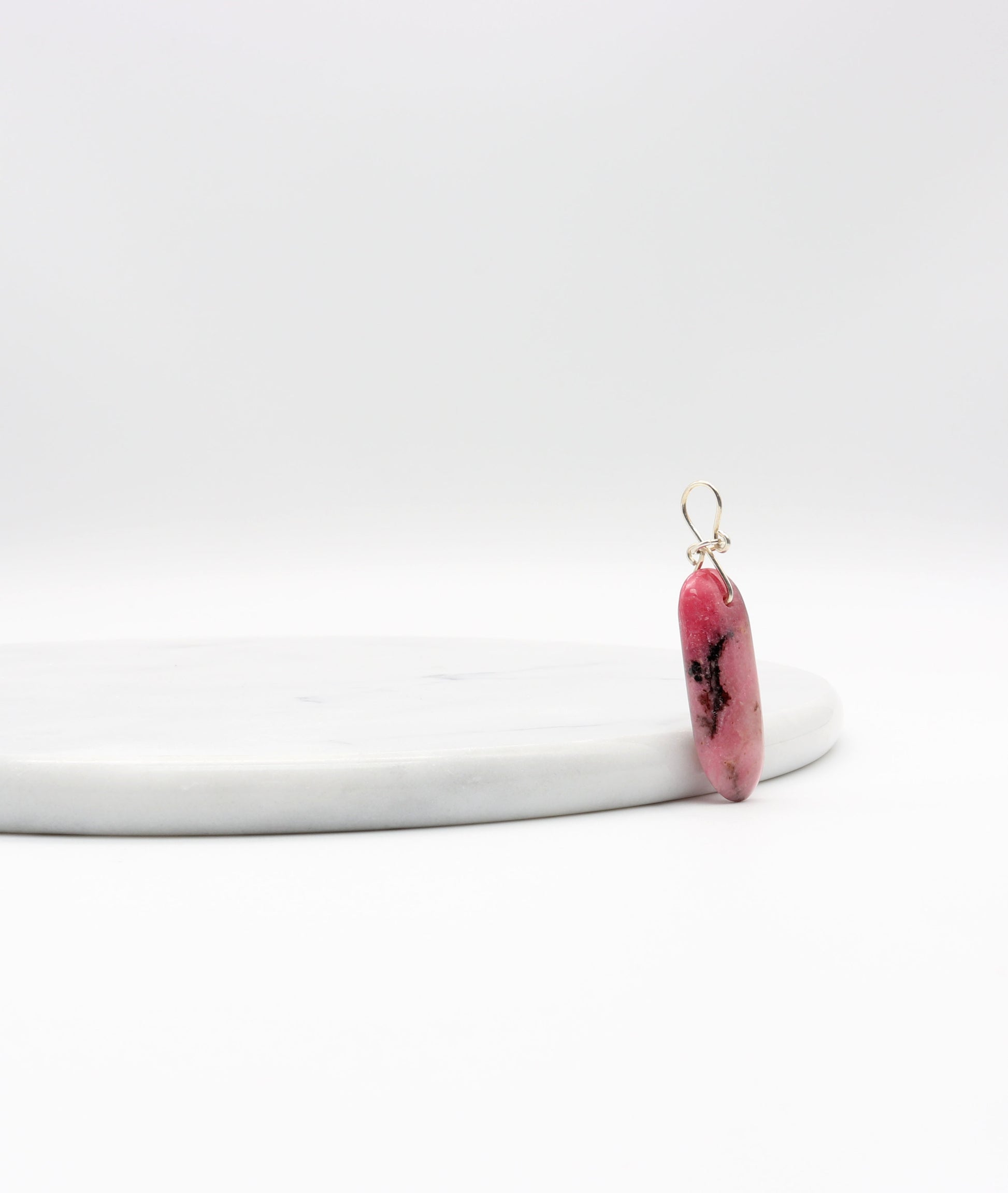 A natural pink Rhodonite crystal pendant with black inclusions, shown next to a white marble slab.
