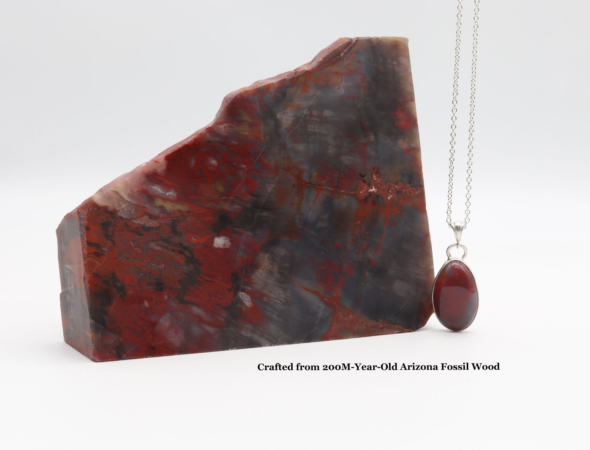 Handcrafted Petrified Rainbow Wood pendant with Argentium 935 Silver bail, next to raw Arizona fossil on light gray surface.  Text overlay crafted from 200m year old Arizona Fossil Wood.