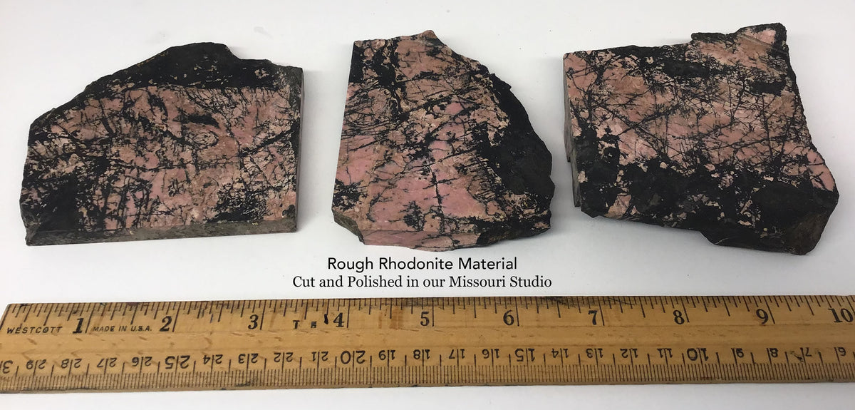Three rough Rhodonite pinkish-black mineral or stone slabs displayed side by side on a white surface, with a wooden ruler along the bottom showing measurements. Text Overlay reads
'Rough Rhodonite Material Cut and Polished in our Missouri Studio'