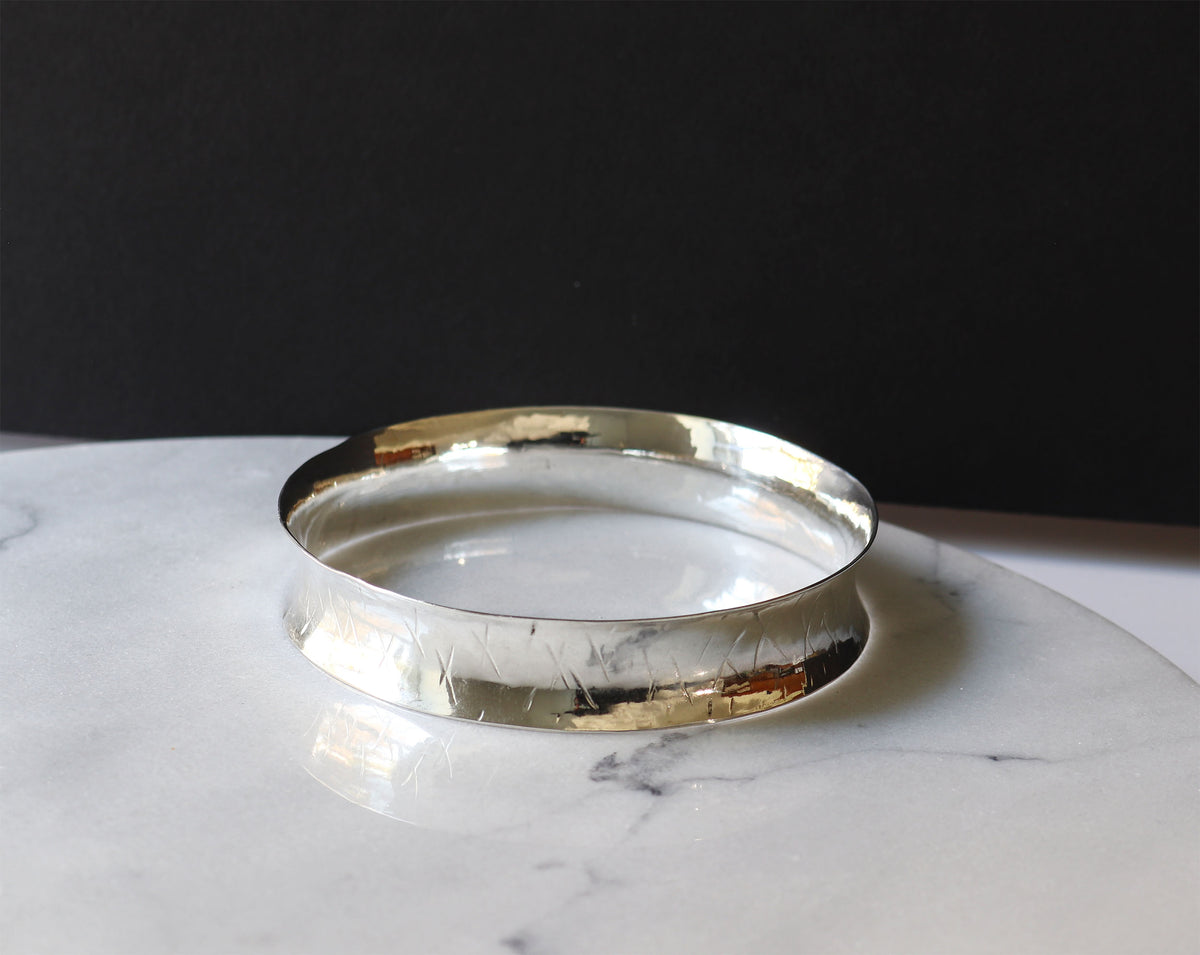 A polished silver-toned metal bangle bracelet with a hammered texture, resting on a white marble surface against a solid black background.
