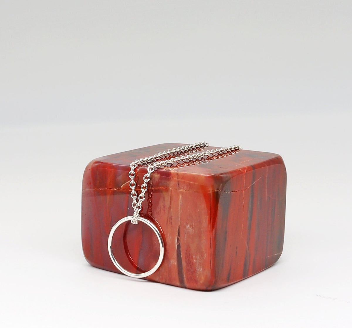 A silver-toned chain necklace with a circular ring pendant draped over a polished petrified rainbow wood red stone square, photographed on a clean white studio background.
