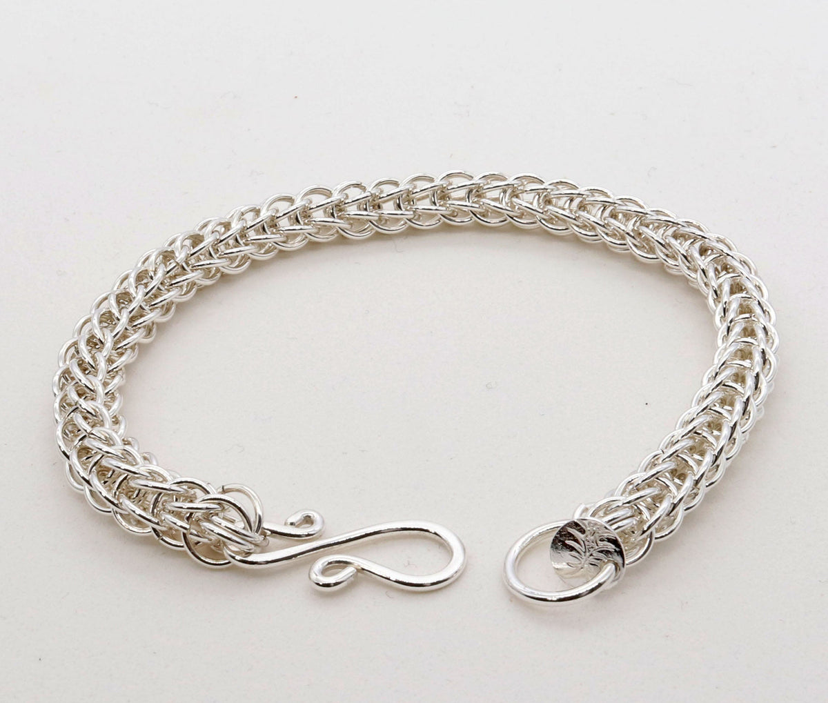 A silver persian weave chain-link bracelet with a unique infinity-shaped clasp and a small circular hallmark charm, displayed on a clean white background.